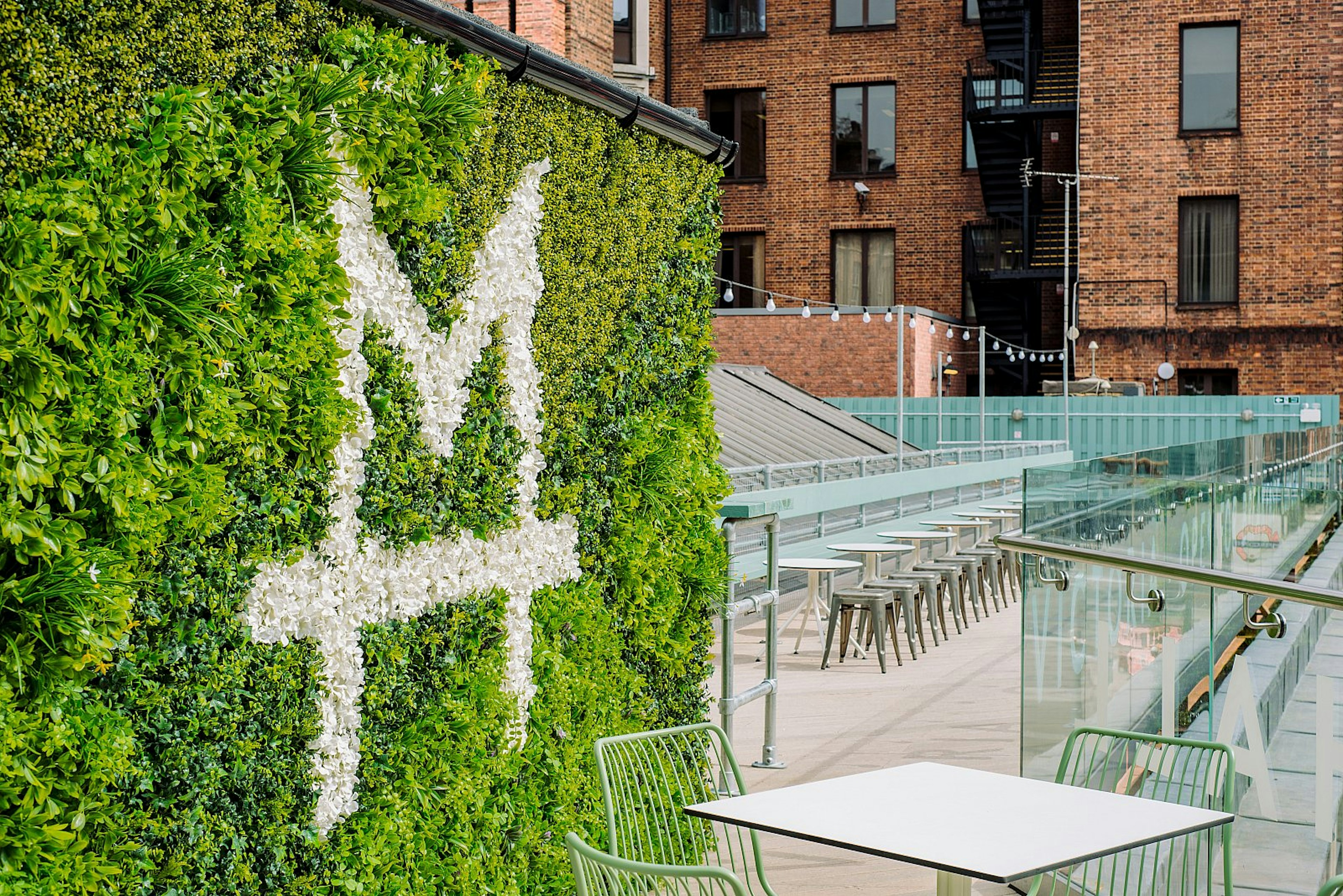 An empty Market Hall Victoria, one of the best rooftop bars in London; in the foreground is a hedge emblazoned with the bar's logo in white, while beyond are white tables and metal stalls; a red-brick building is beyond.