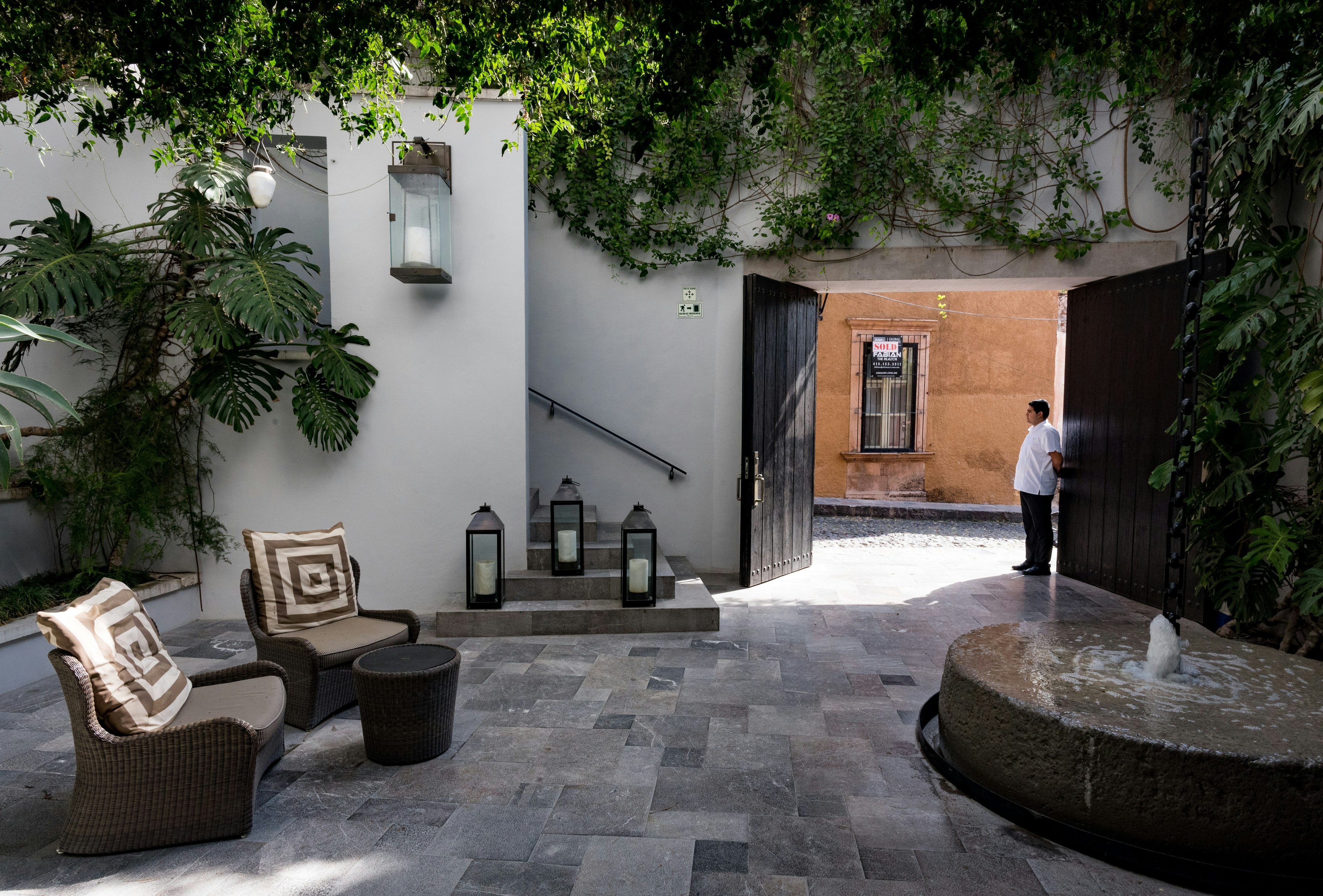 A hotel employee stands behind a stone fountain in the airy, greenery-draped courtyard of a hotel; the terracotta-colored walls of the street beyond are visible behind him