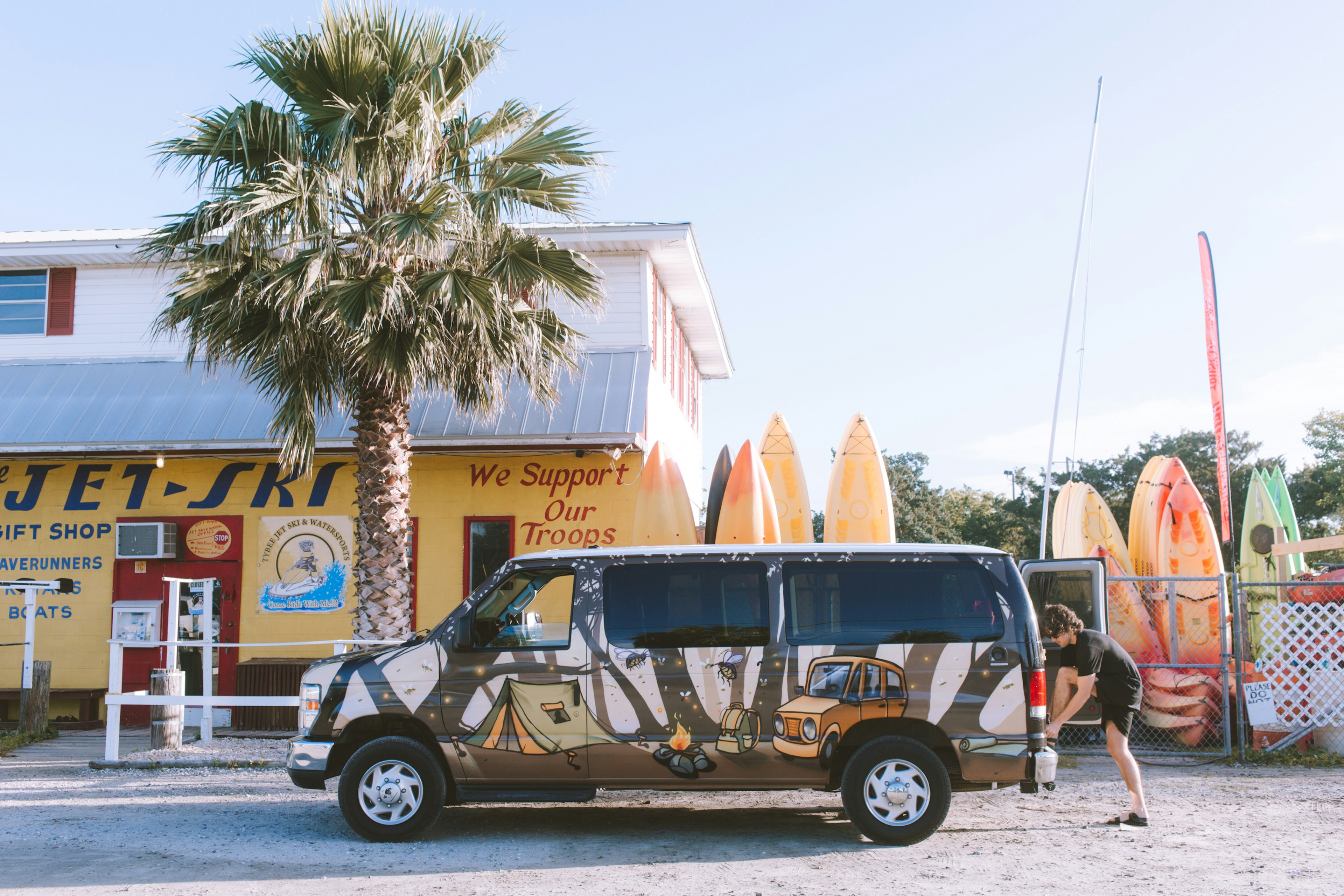 A man ties his shoe on the back of a painted campervan parked in front of a surf shop