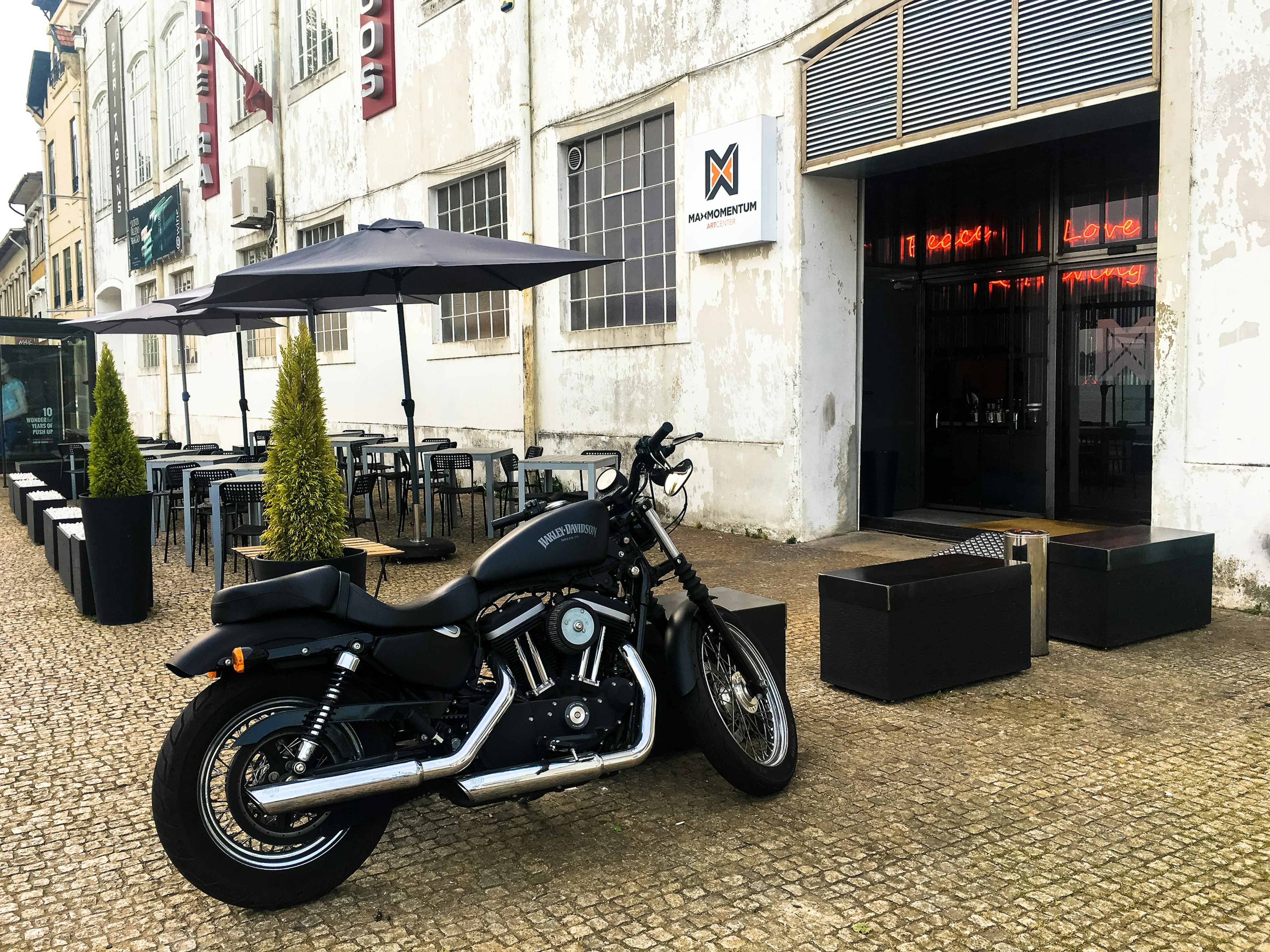 The minimalist entrance to the grey building housing Maxmomentum has been dressed up with potted topiary, tables, chairs and black parasols. A motorbike is propped up outside.