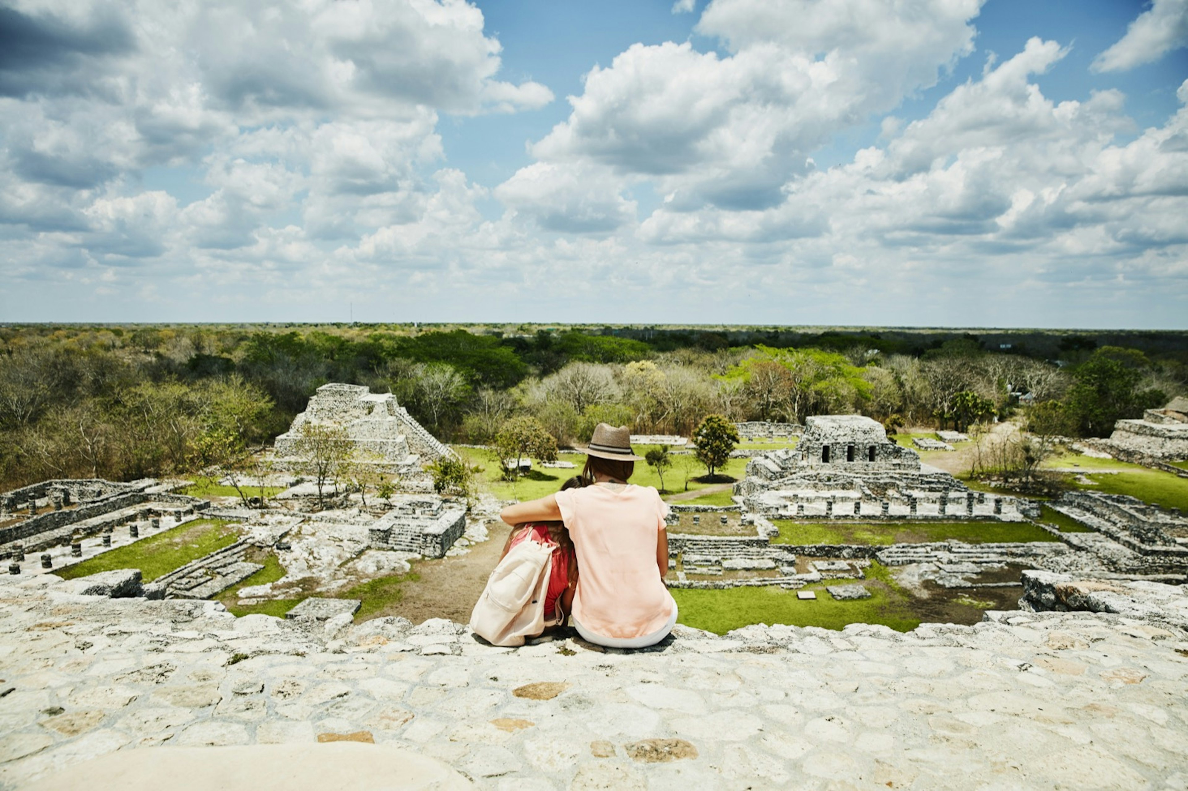 A woman and young girl embrace while sitting on a stone surface and looking out over the ruins of Mayapan