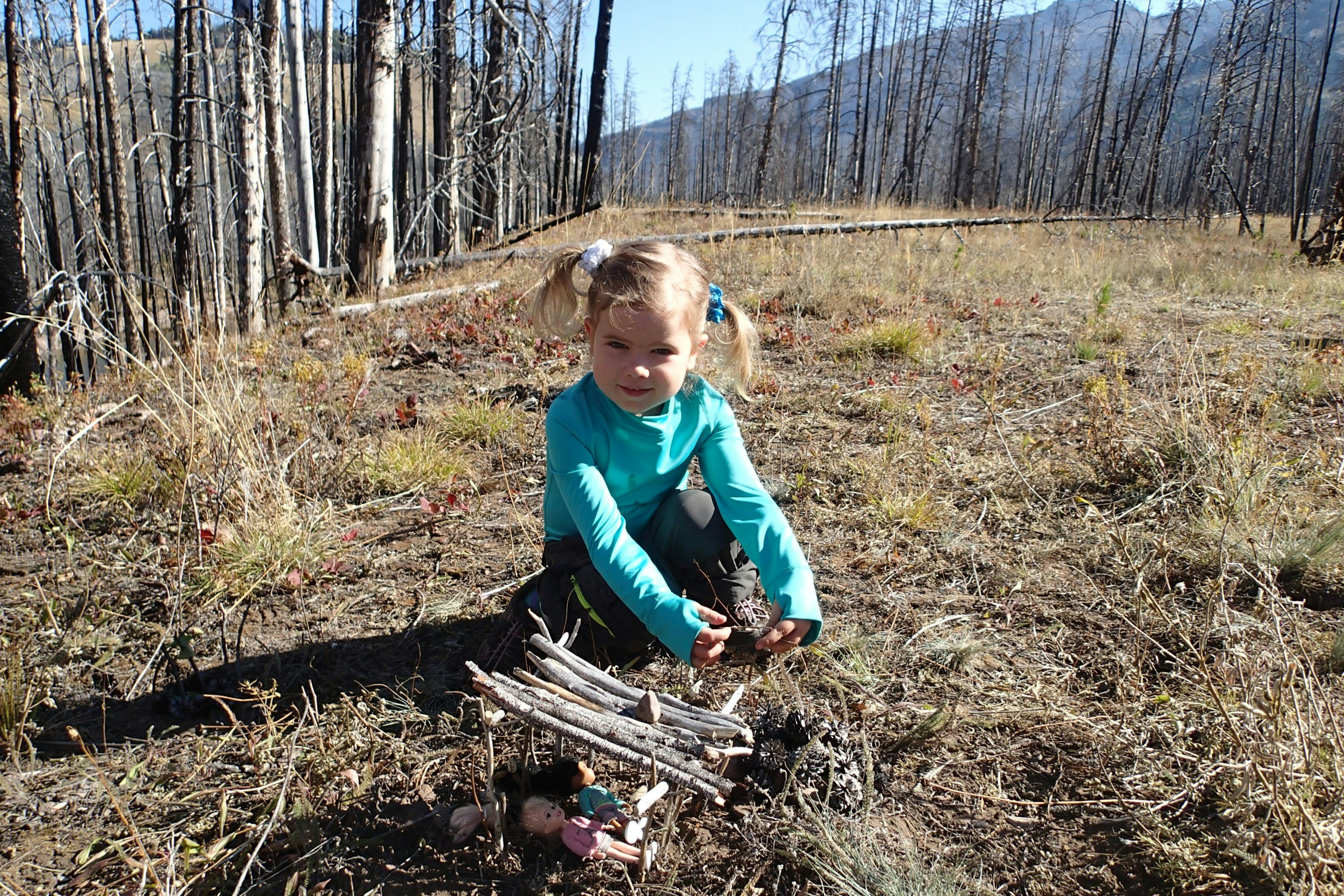 A young girl poses next to a stick shelter, covering her two small baby dolls; Project Remote