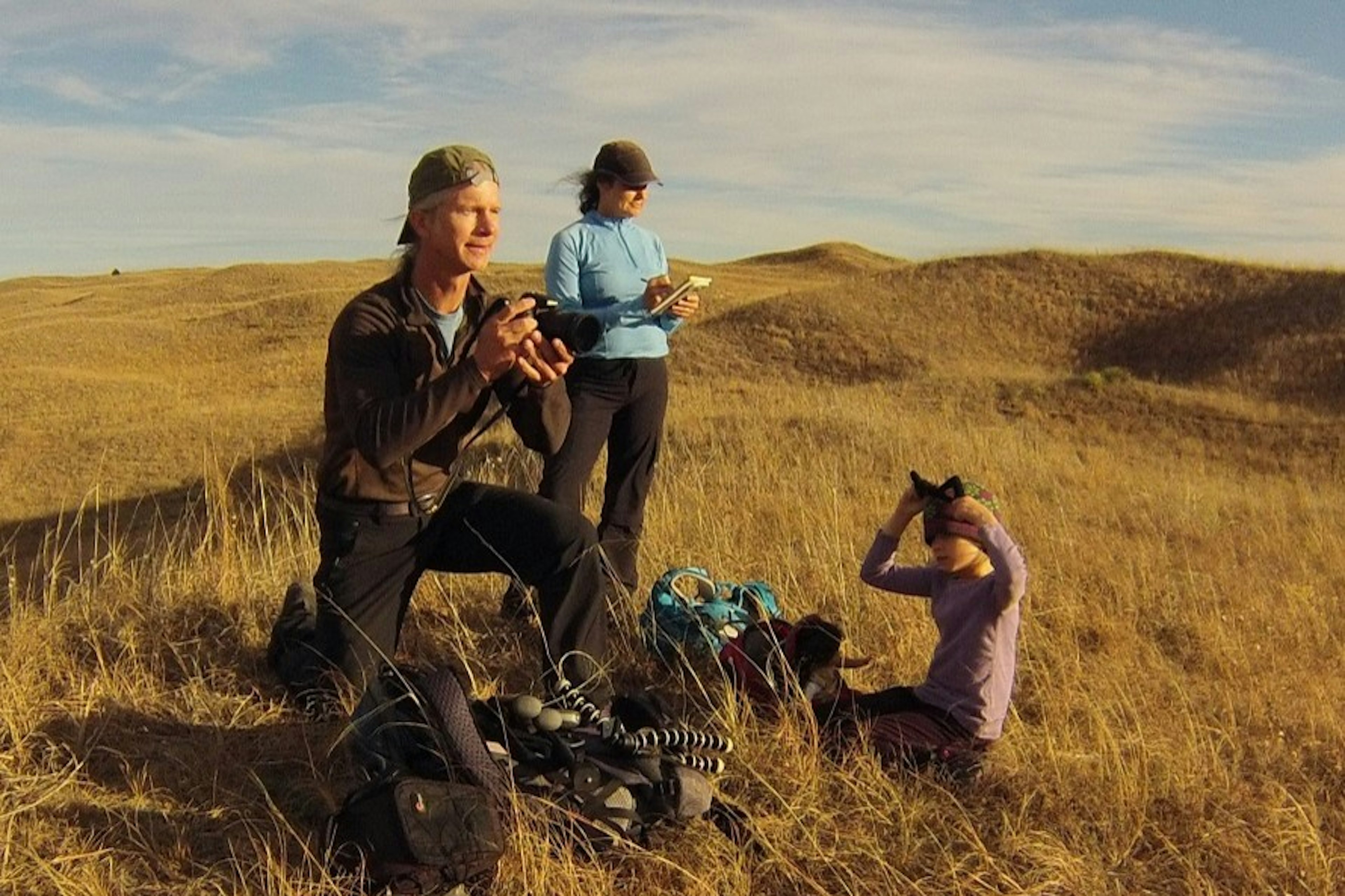 A man and a woman look out over a golden field of grass as their young daughter plays at their feet; Project Remote