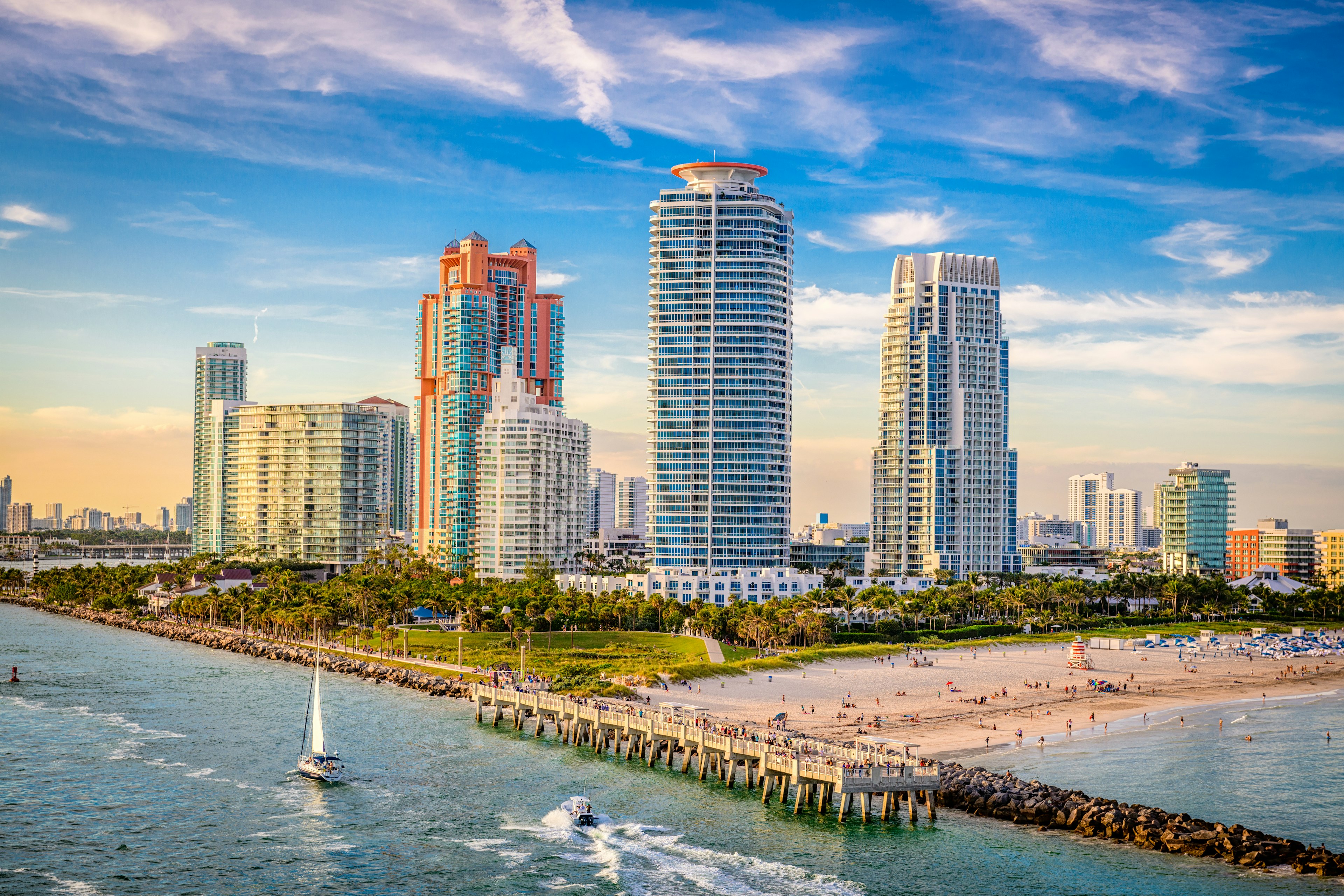 Aerial view of the South Beach skyline dotted with high-rise buildings. In the foreground, a sailboat and a speed boat sail past a wooden pier which separates the beach from the ocean.