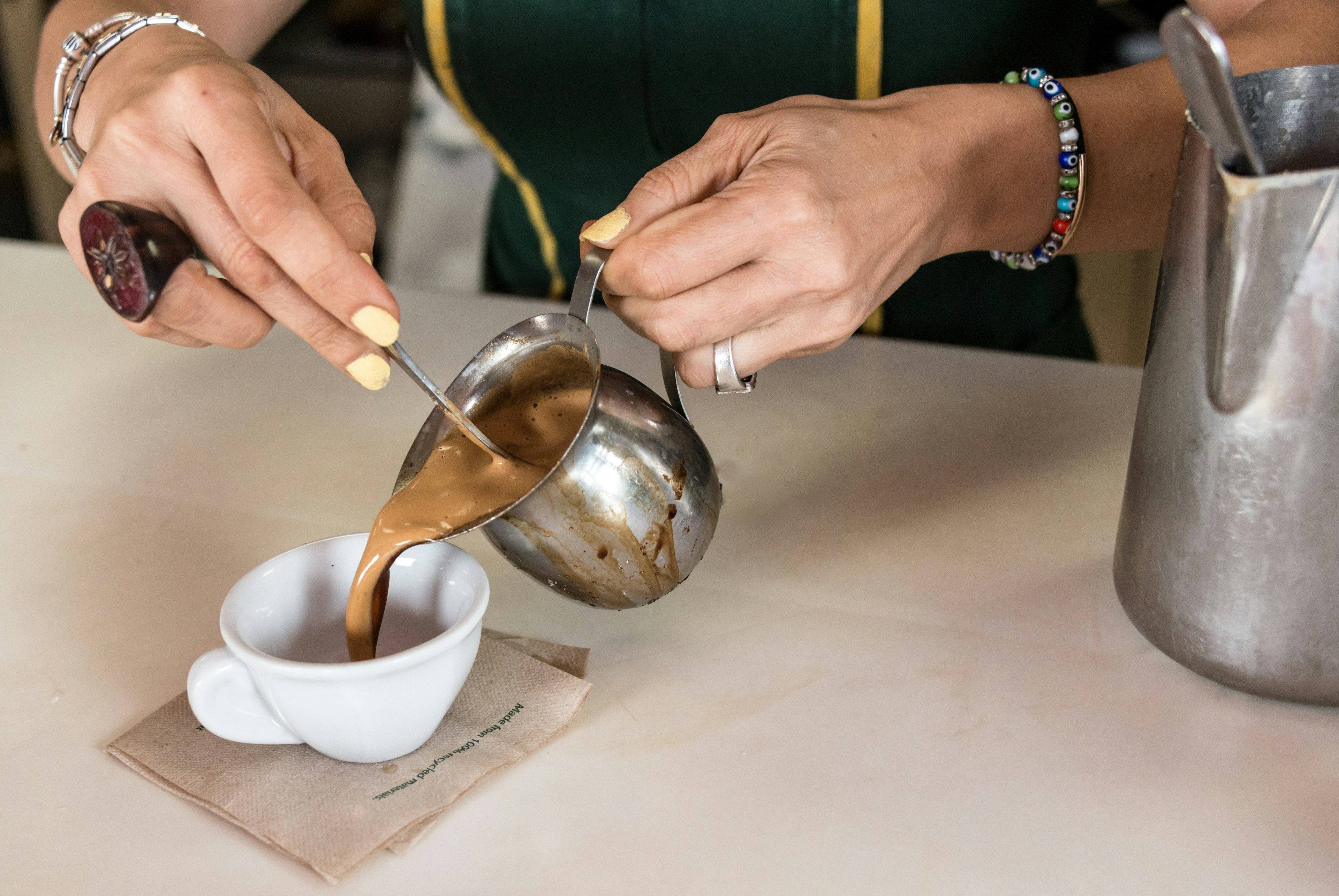 Someone is pouring a light brown Cuban coffee out of a metal jug into a small white cup