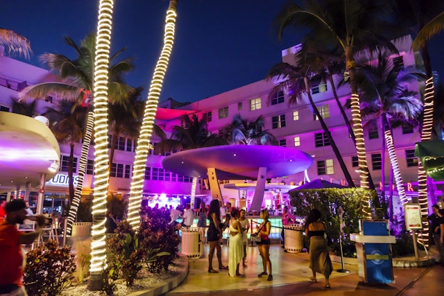 Young women chat in front of a Miami outdoor nightclub dominated by a palm tree wrapped in lights