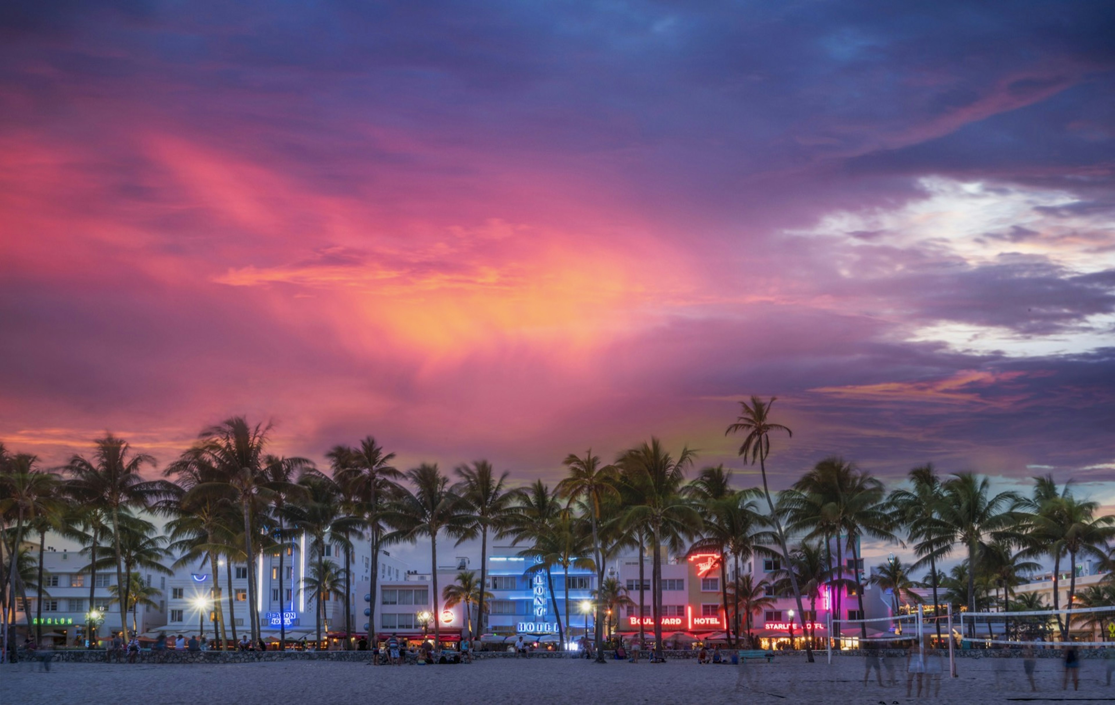 The strip in Miami with neon lit buildings, palm trees and a pink sky behind