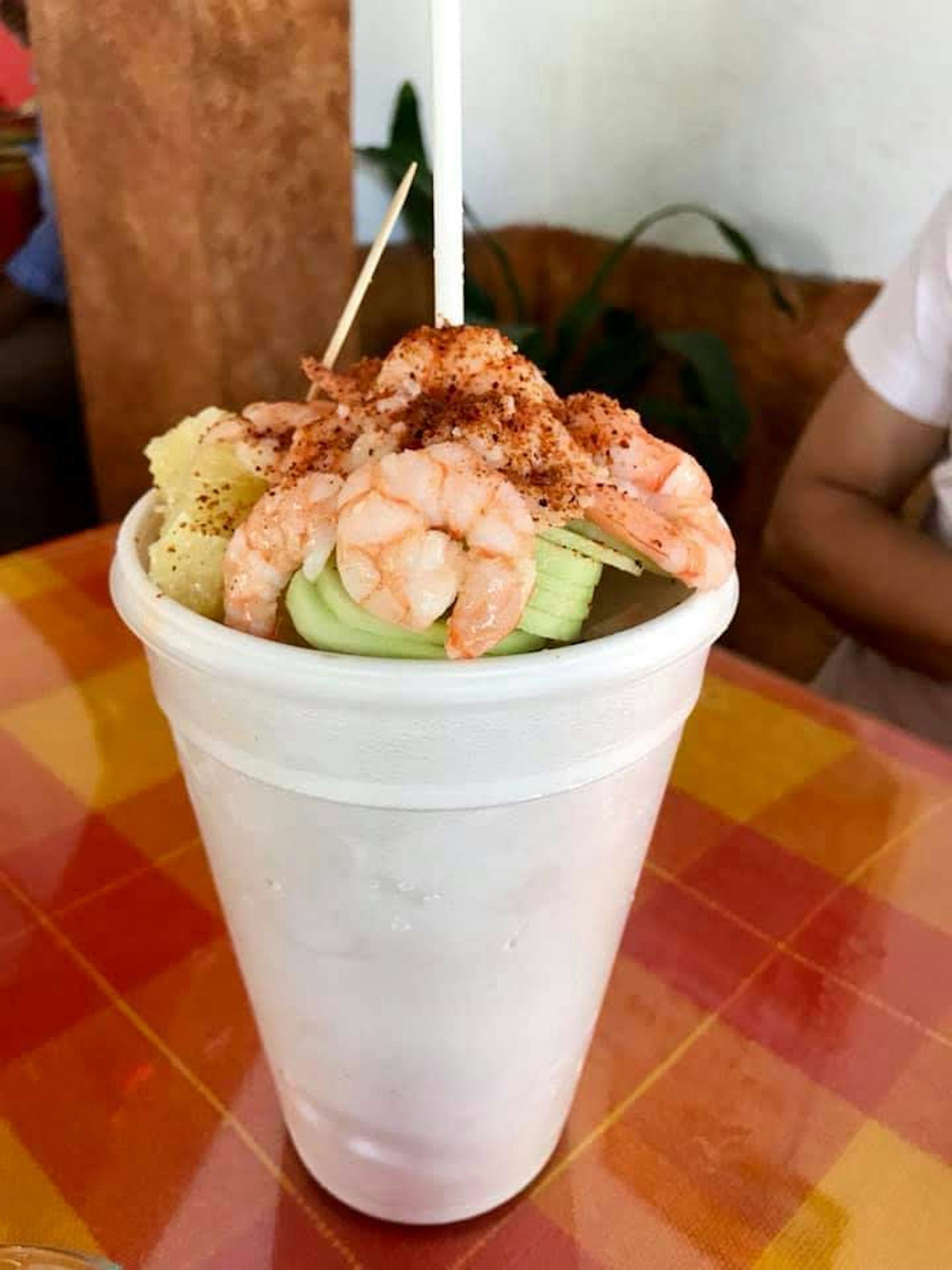 A tall, white styrofoam cup filled with Mexican beer and topped with shrimp, cucumbers, pineapple and sprinkled with spicy powder sits on a plastic checkered table cloth