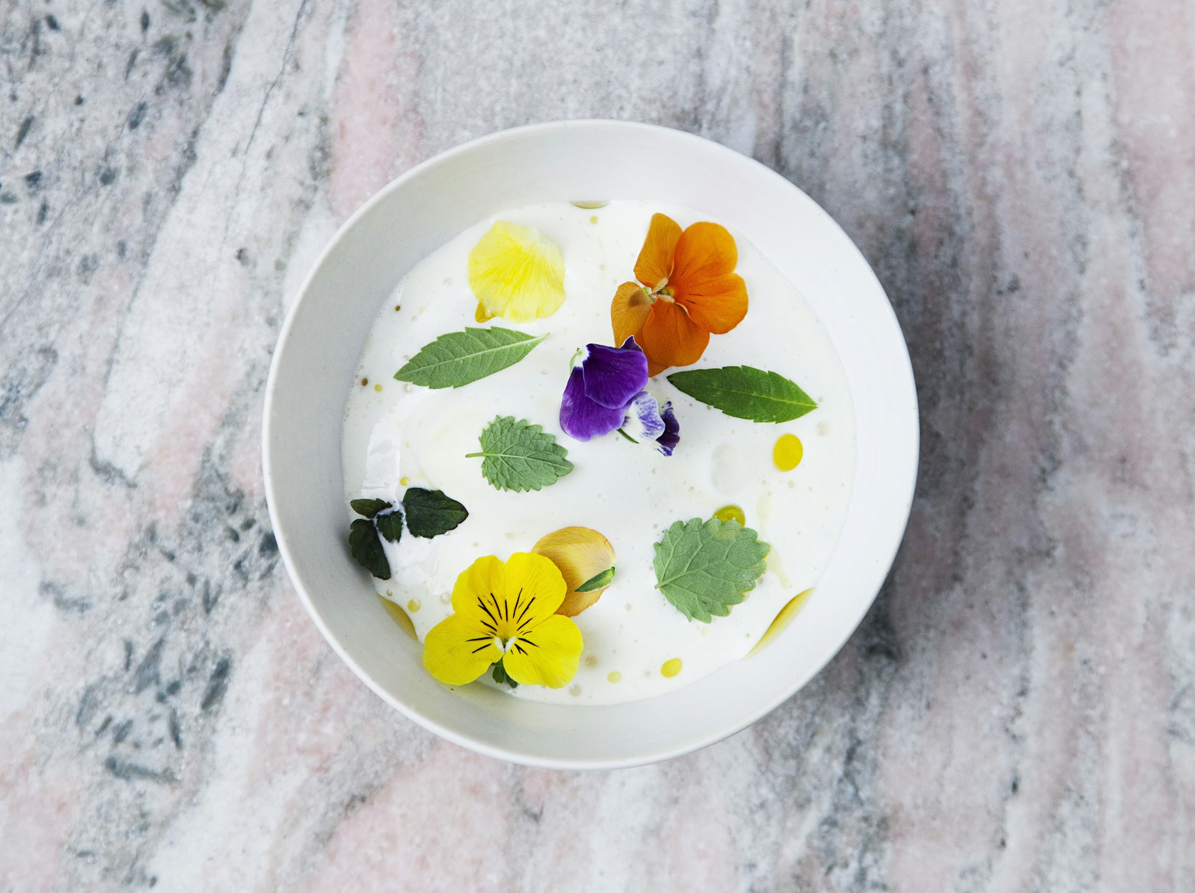 A white bowl on a marble table is filled with a white soup, with bright orange, purple and yellow flowers floating atop it.