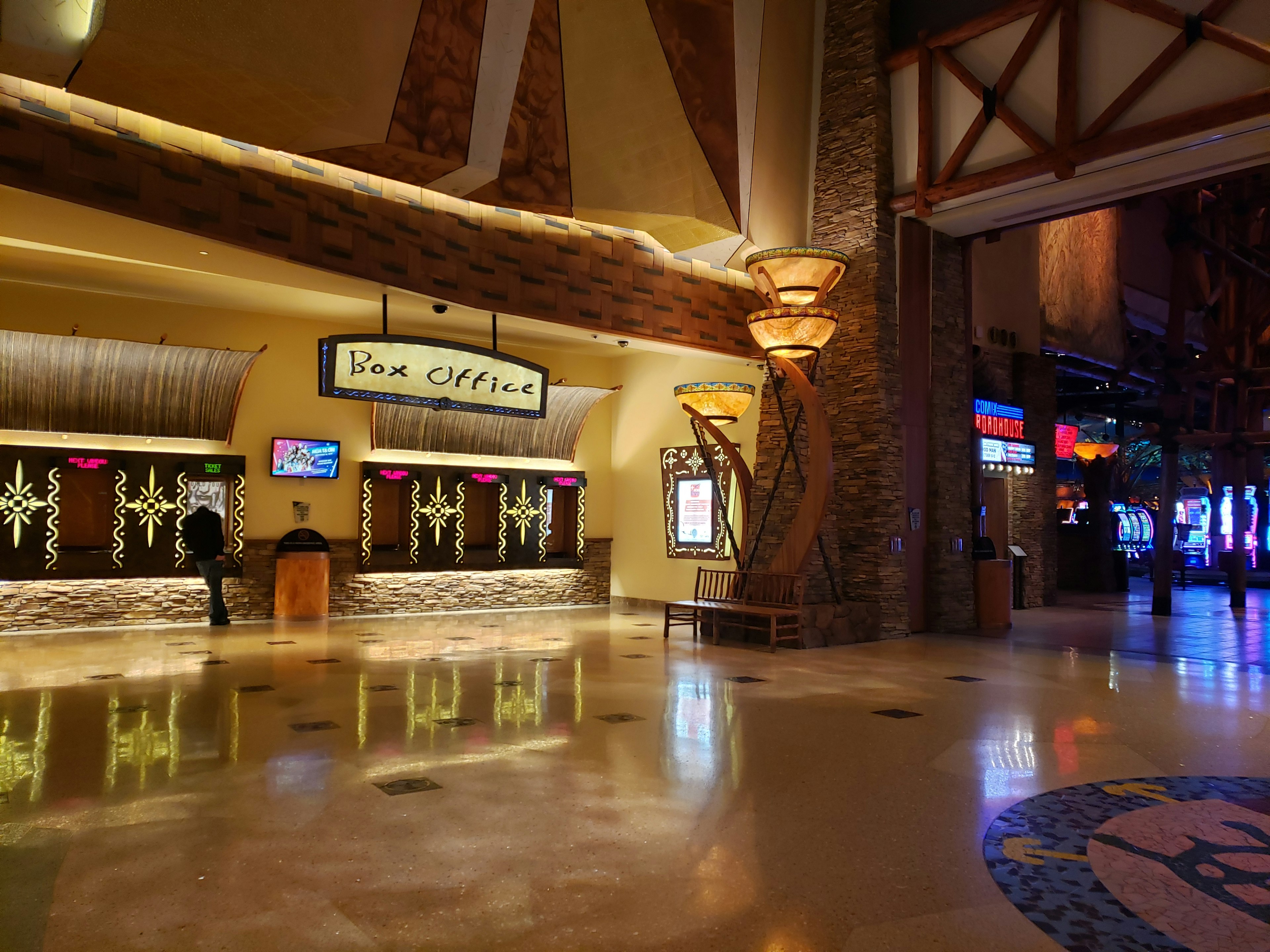 The golden-hewed lobby of the Arena at Mohgen Sun Casino is decorated with a flagstone column, contemporary stained glass light fixtures, curved warm wood, and abstract modernist forms in yellow adn brown hues. A large sign says Box Office in a rustic font. In the bottom right corner of the frame, a circular blue mosaic in the floor can be seen. In the distance, glowing signs from the Comix Roadhouse and arcades shine.