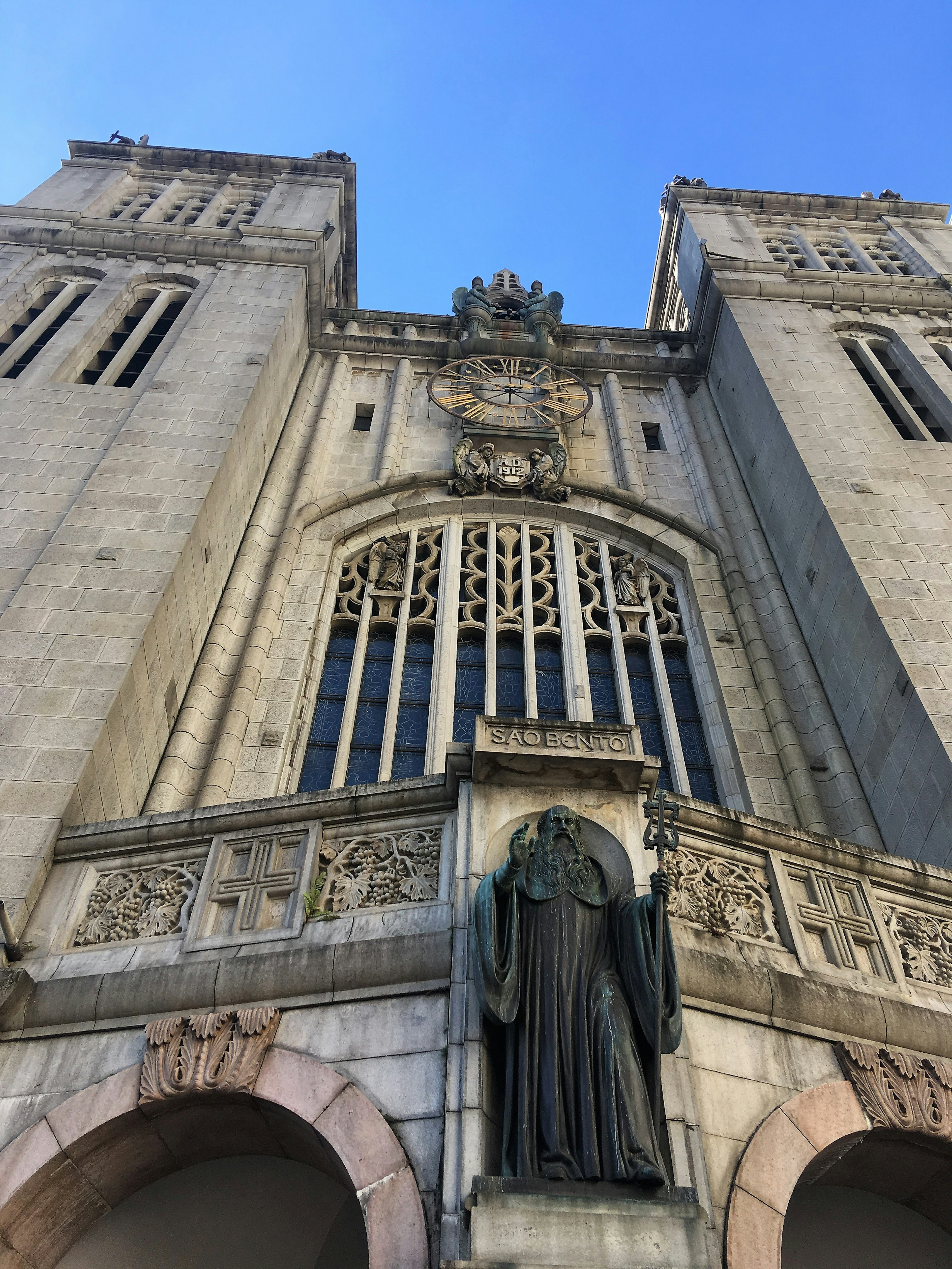 Looking up at the grey stone neo-Gothic facade of Mosteiro São Bento, with two towers flanking a statue of Christ, a tall stained glass window and a clock.