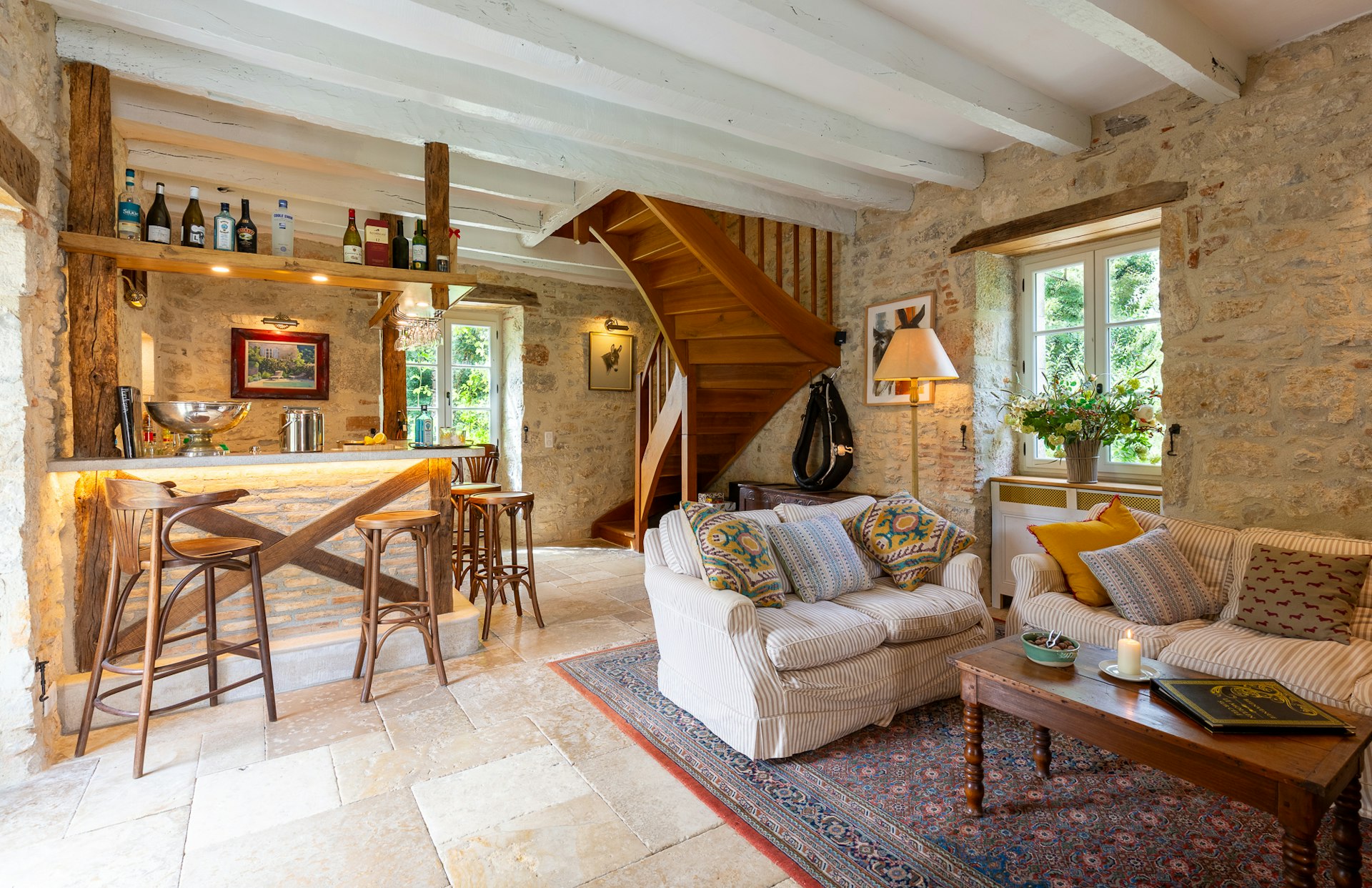 A lounge room with stone walls, sofas and a corner bar. A wooden spiral staircase is in the corner of the room