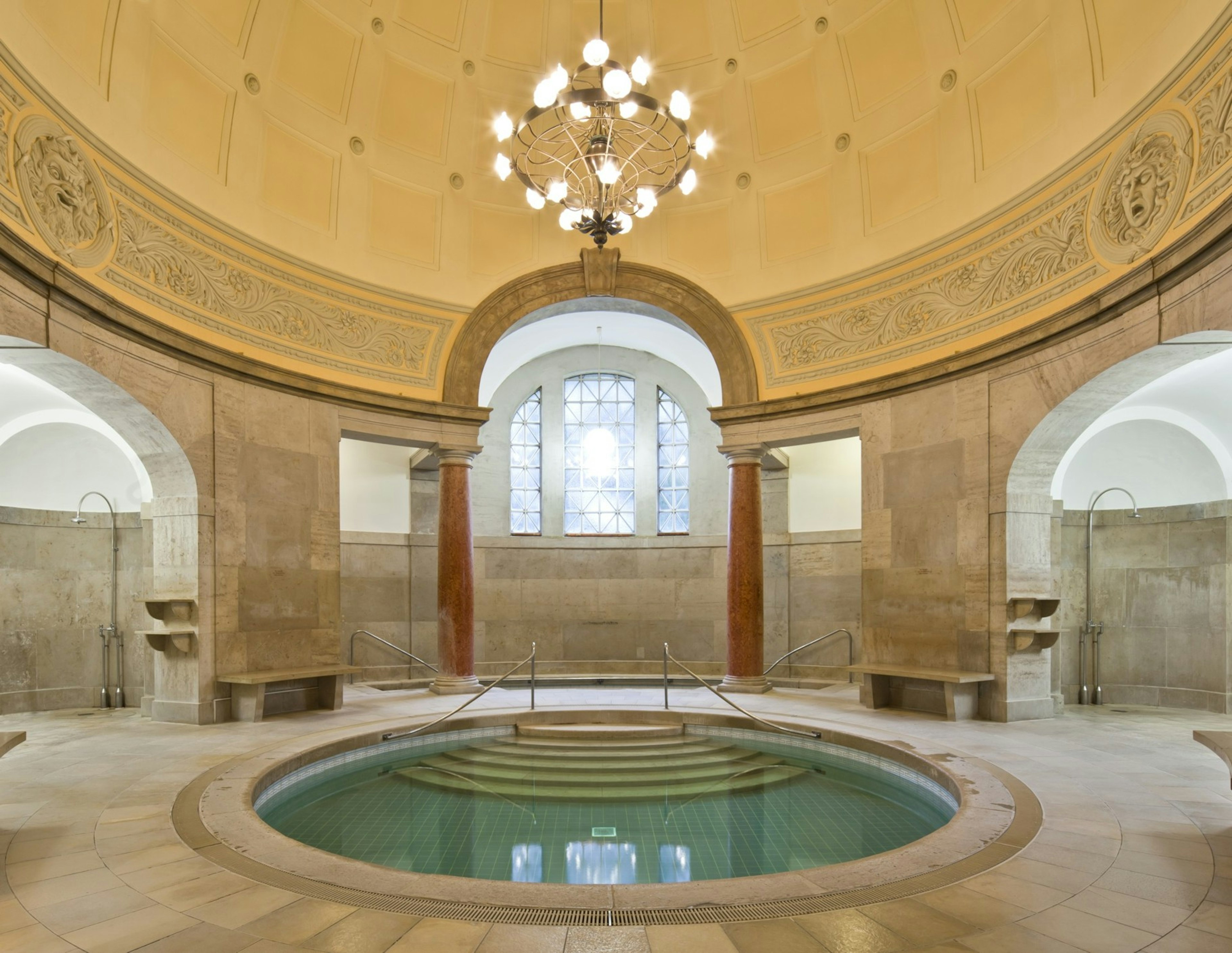 A blue circular pool in a marbled hall with a high-domed ceiling and chandelier in. There are known as 'hot corners' in Müller’sche Volksbad, a beautiful bathhouse in Munich.