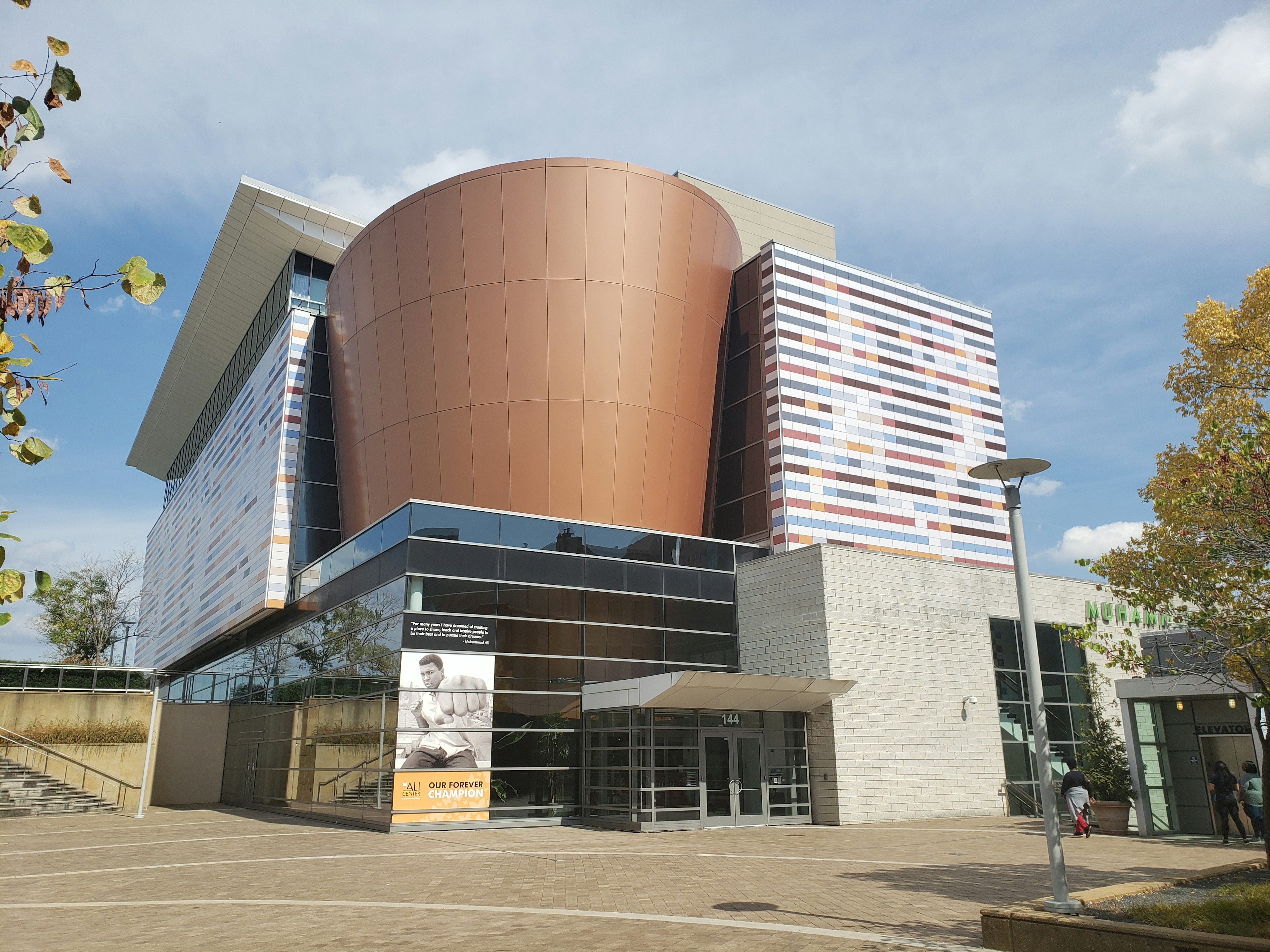 The exterior facade of the Muhammed Ali Center in Louisville, Kentucky is made up of several modernist forms, including a large bronze-colored curved tower over a rectangular glass lobby, surrounded by rectangular walls with a muted blue, red, and orange pattern that looks almost pixelated