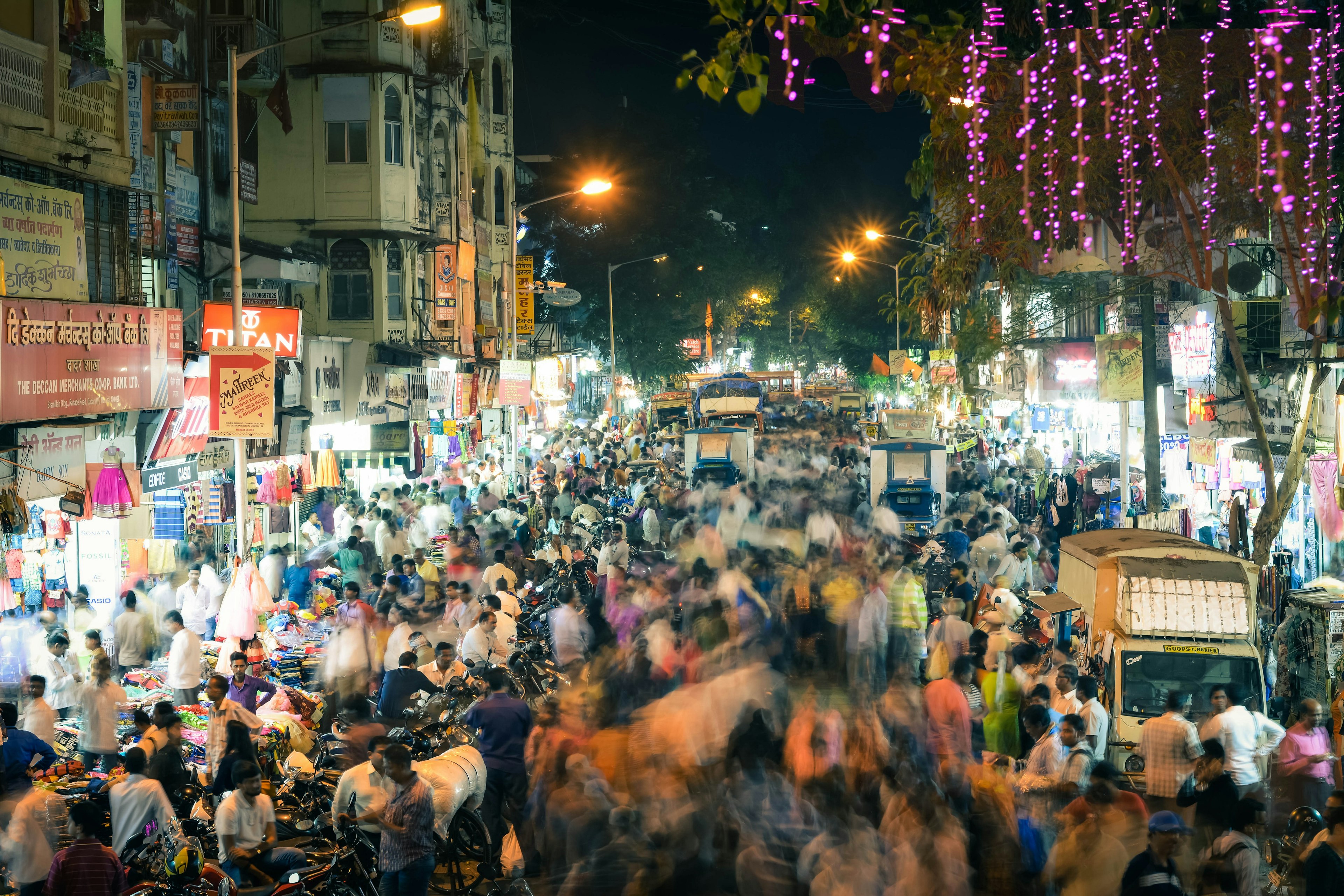 A view of a very busy road in Mumbai, flanked on either side by makeshift stalls and shops. The crowds walking through the street are blurred slightly, giving the appearance that they are in motion.