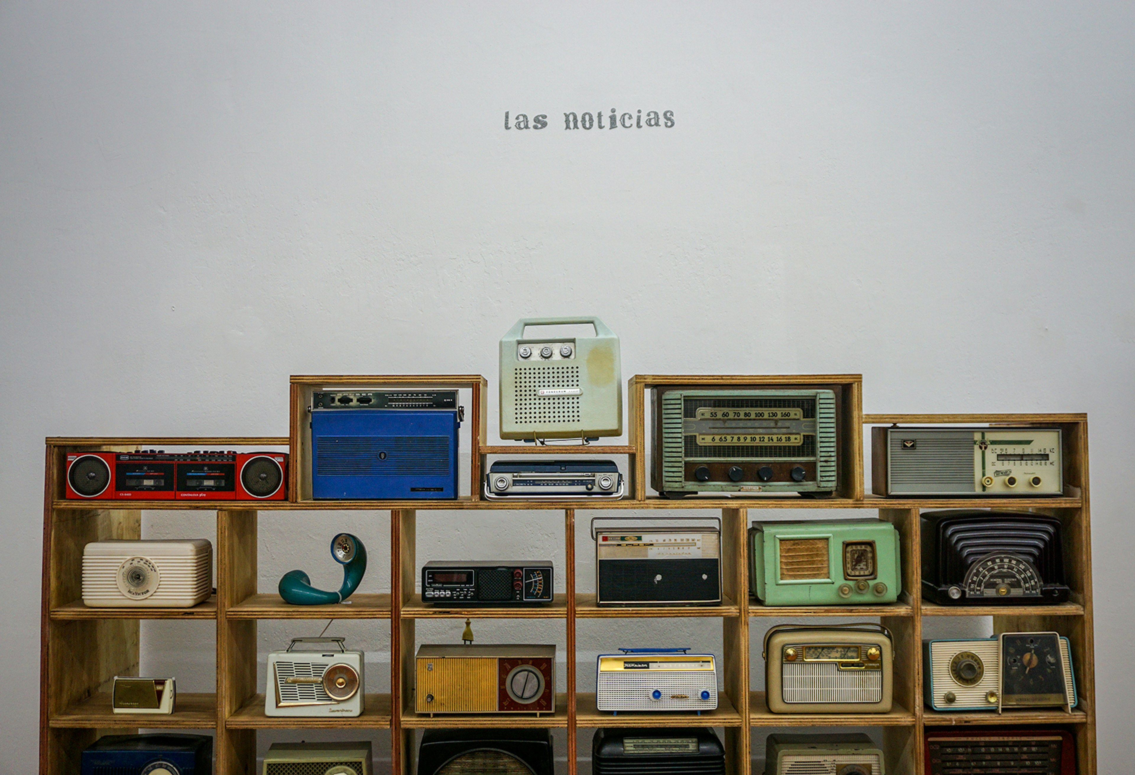 Old radios stacked in a mid-century style shelf against a blank white wall, with