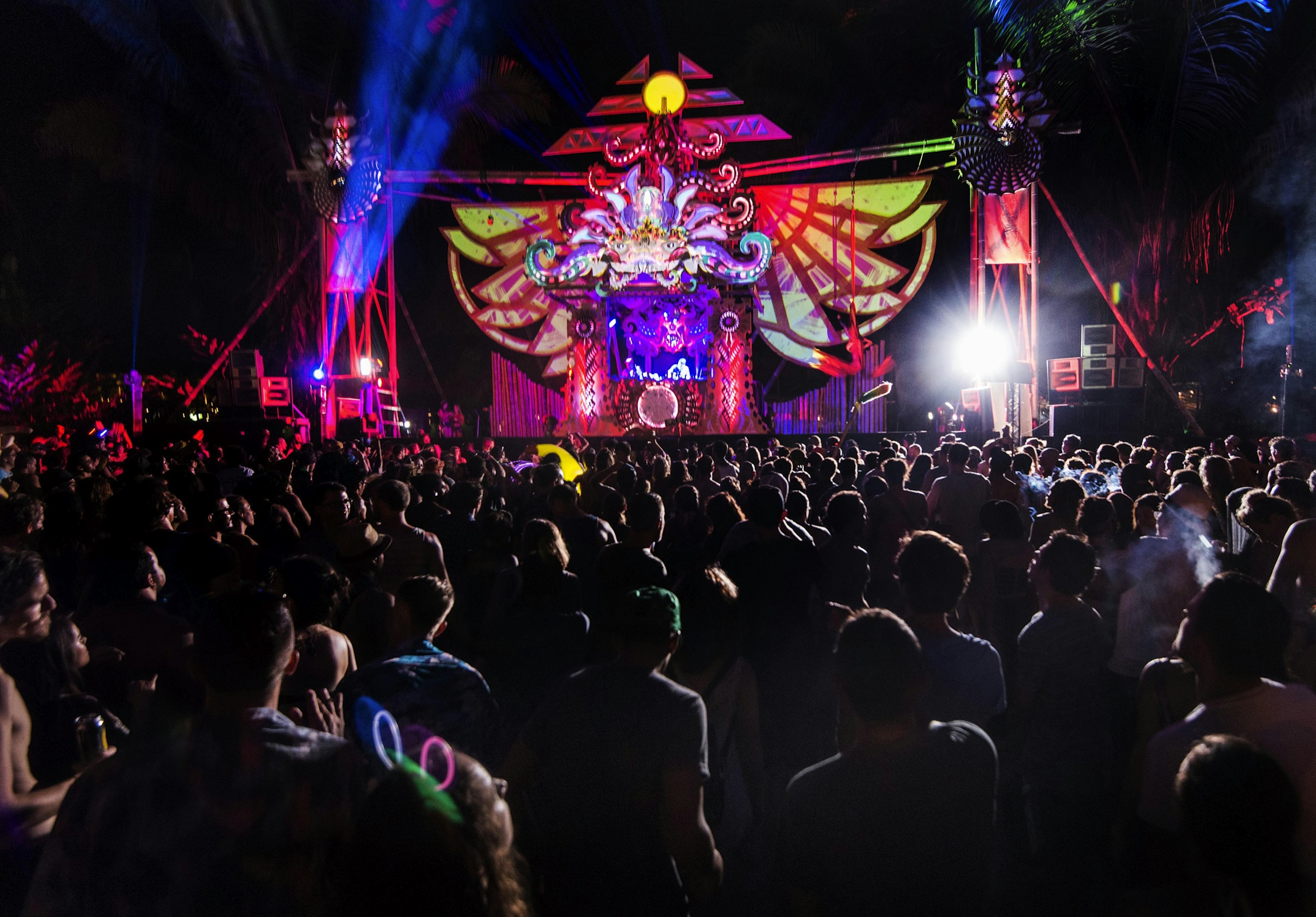 A festival crowd gathers in front of a music stage lit with neon lights and laserbeams.