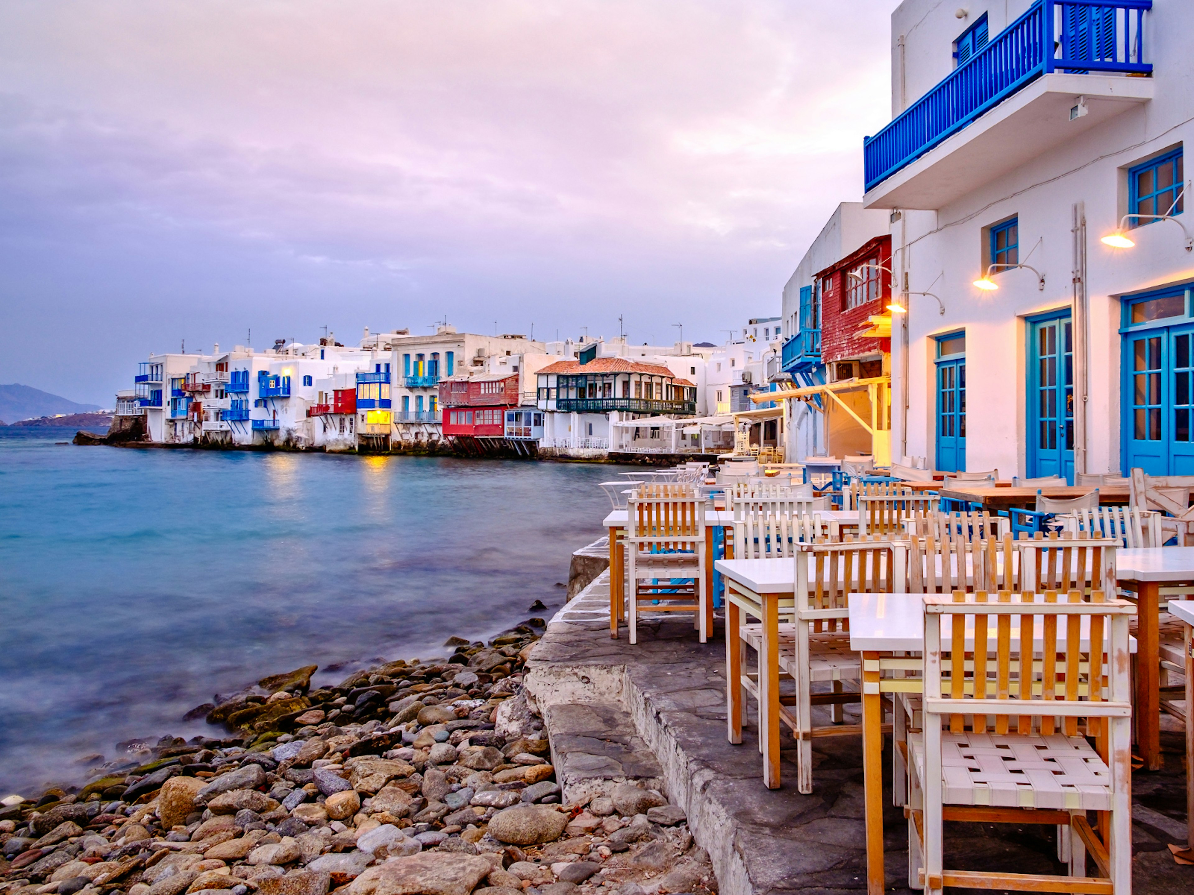 White buildings with colourful balconies and windows line up to the shore. In the foreground, a restaurant area is laid out with tables and chairs