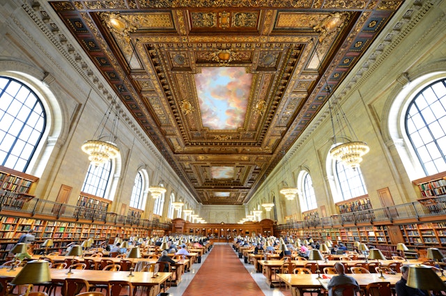 Interior shot of the expansive Rose Main Reading room at the New York Public Library. There are multiple wooden tables and a very ornate ceiling with large chandeliers.