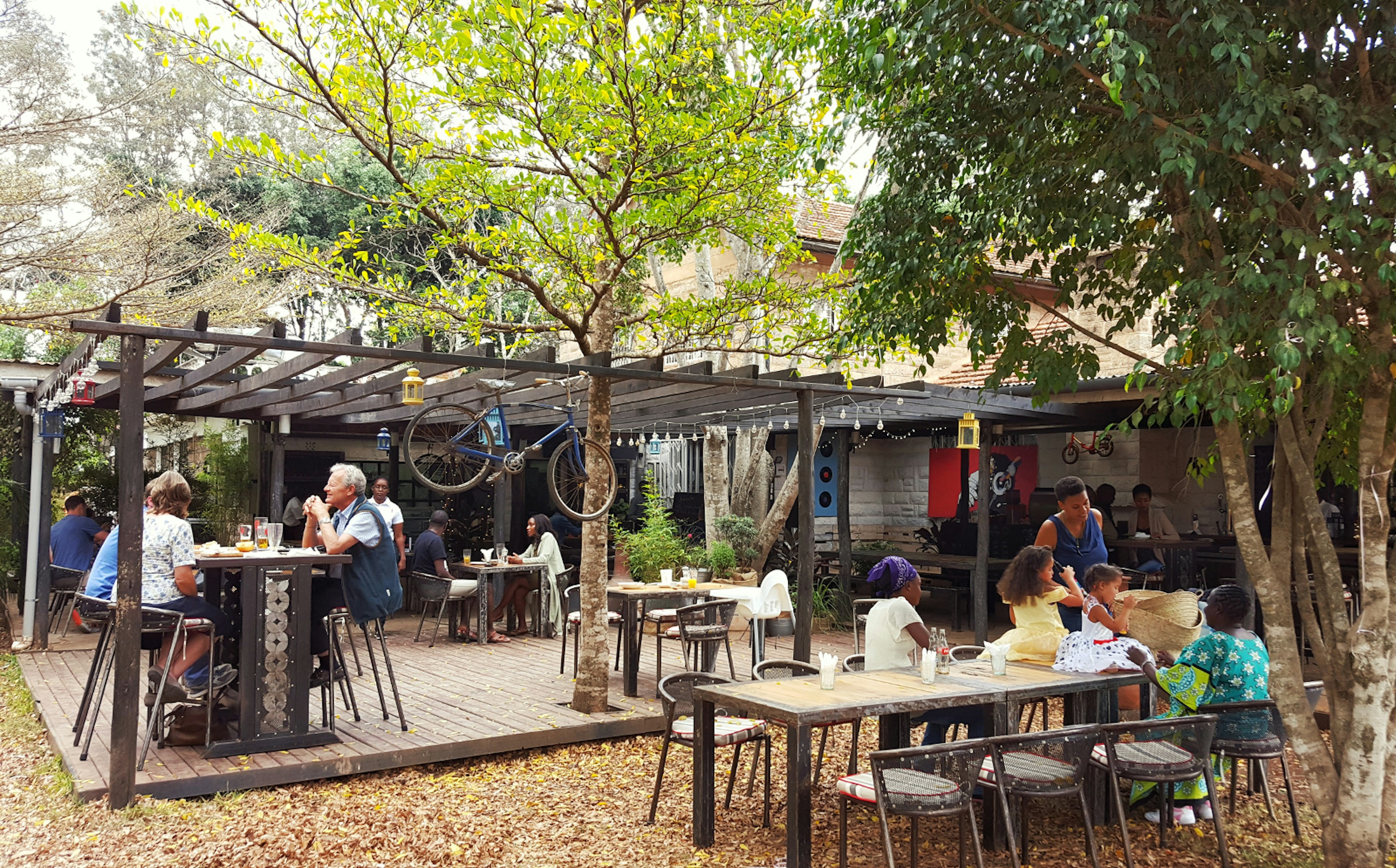 Leafy trees shade a large outdoor seating area that is dotted with tables, some on a wooden deck, others on the leaf-covered grass. A bike and candle-lamps hang from an open-slatted roof structure over the deck. Patrons chat, eat and drink, with a family taking over a large table, complete with children (three- and five-year-olds sitting atop the table itself) © Clementine Logan / ϰϲʿ¼