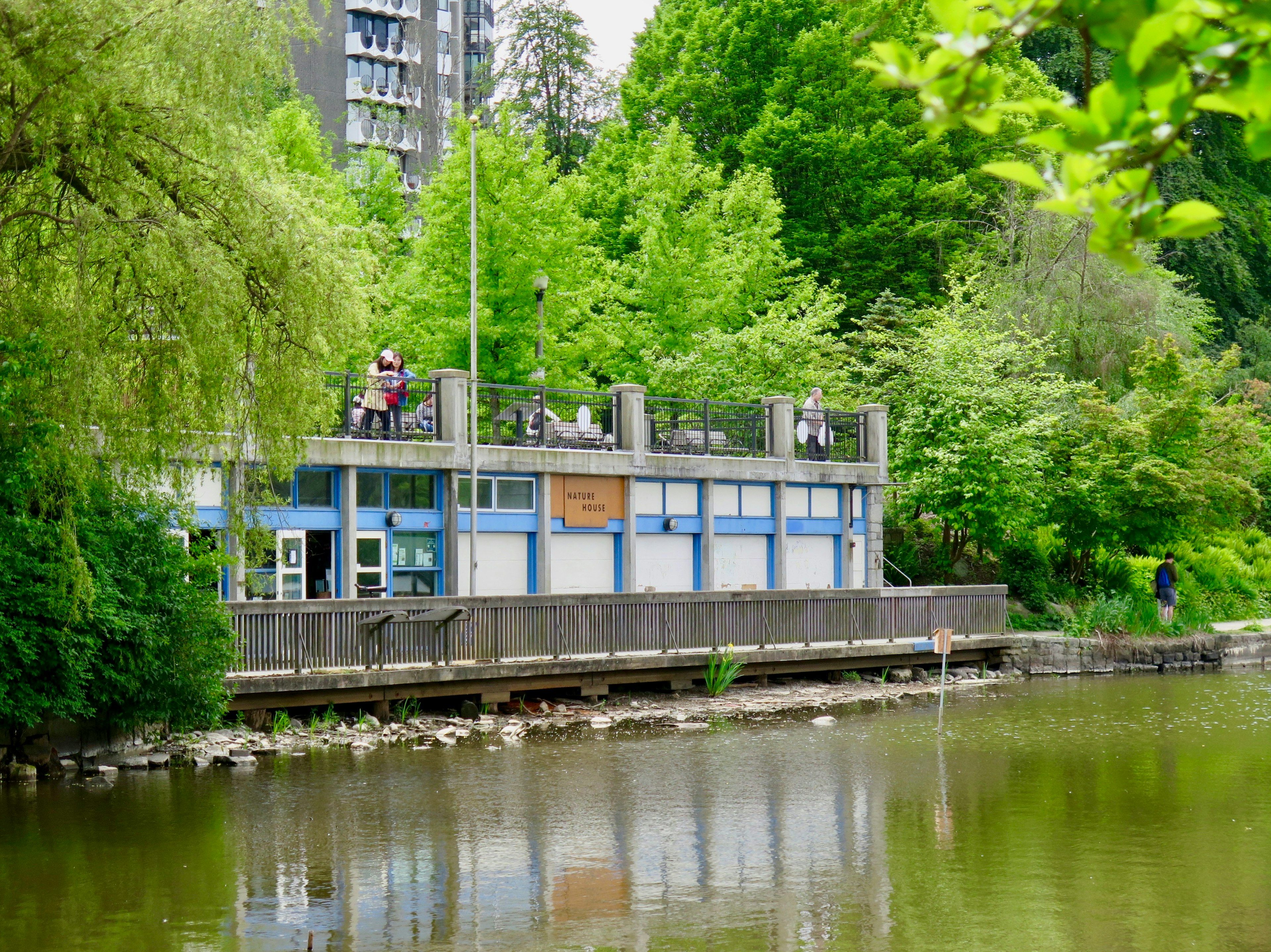 People stand on top of a boxy building set over a marshy water area with a sign that reads Nature House in Vancouver