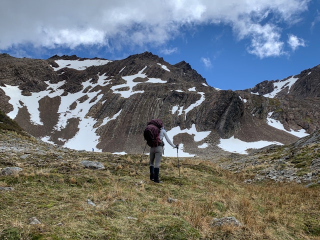 Hiker with backpack and hiking poles stands on a grassy hill looking at snow-streaked mountains