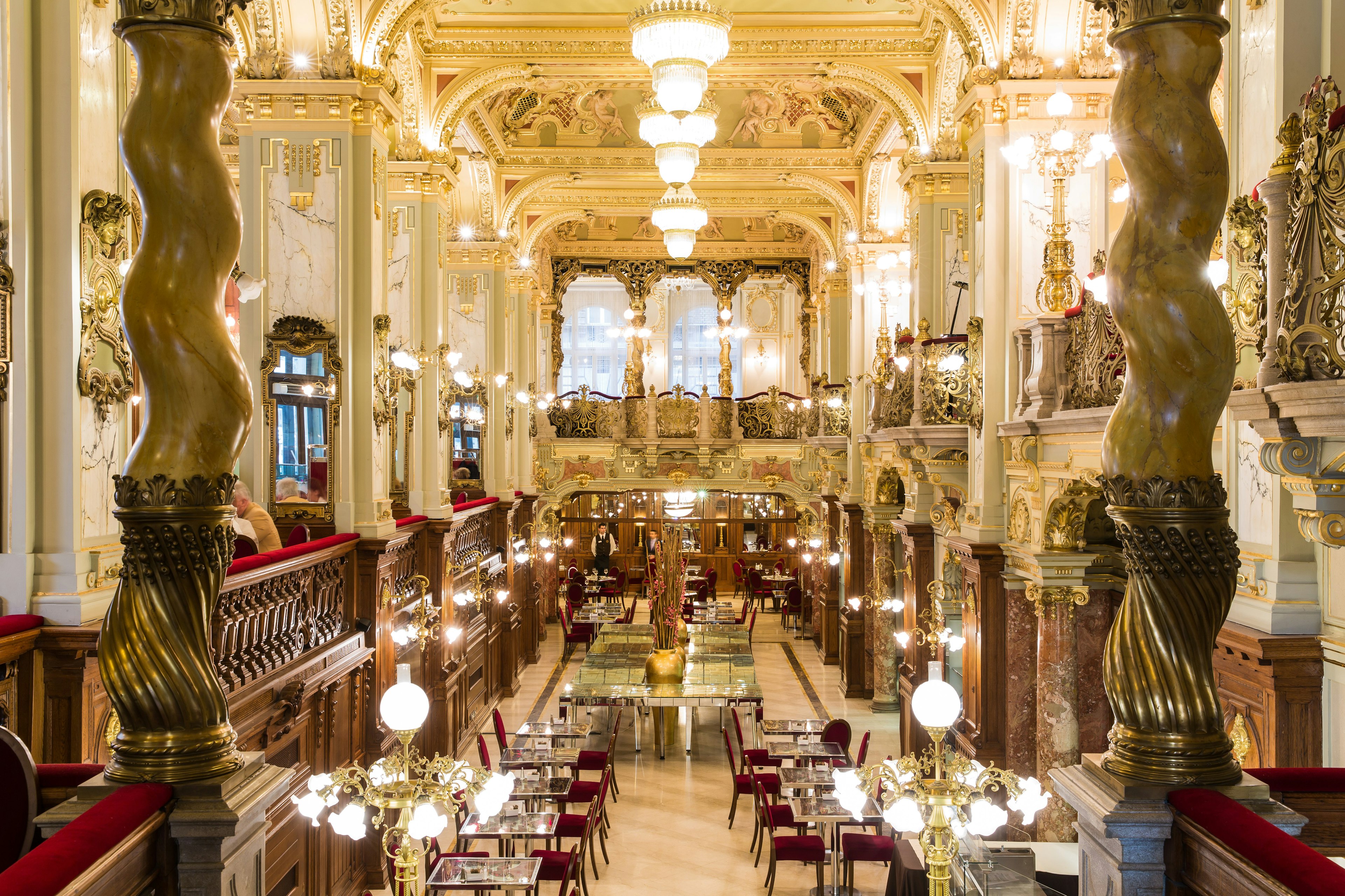 The wildly ornate interior of the New York Café in Budapest. There are marble columns, golden light fixtures and plush crimson chairs