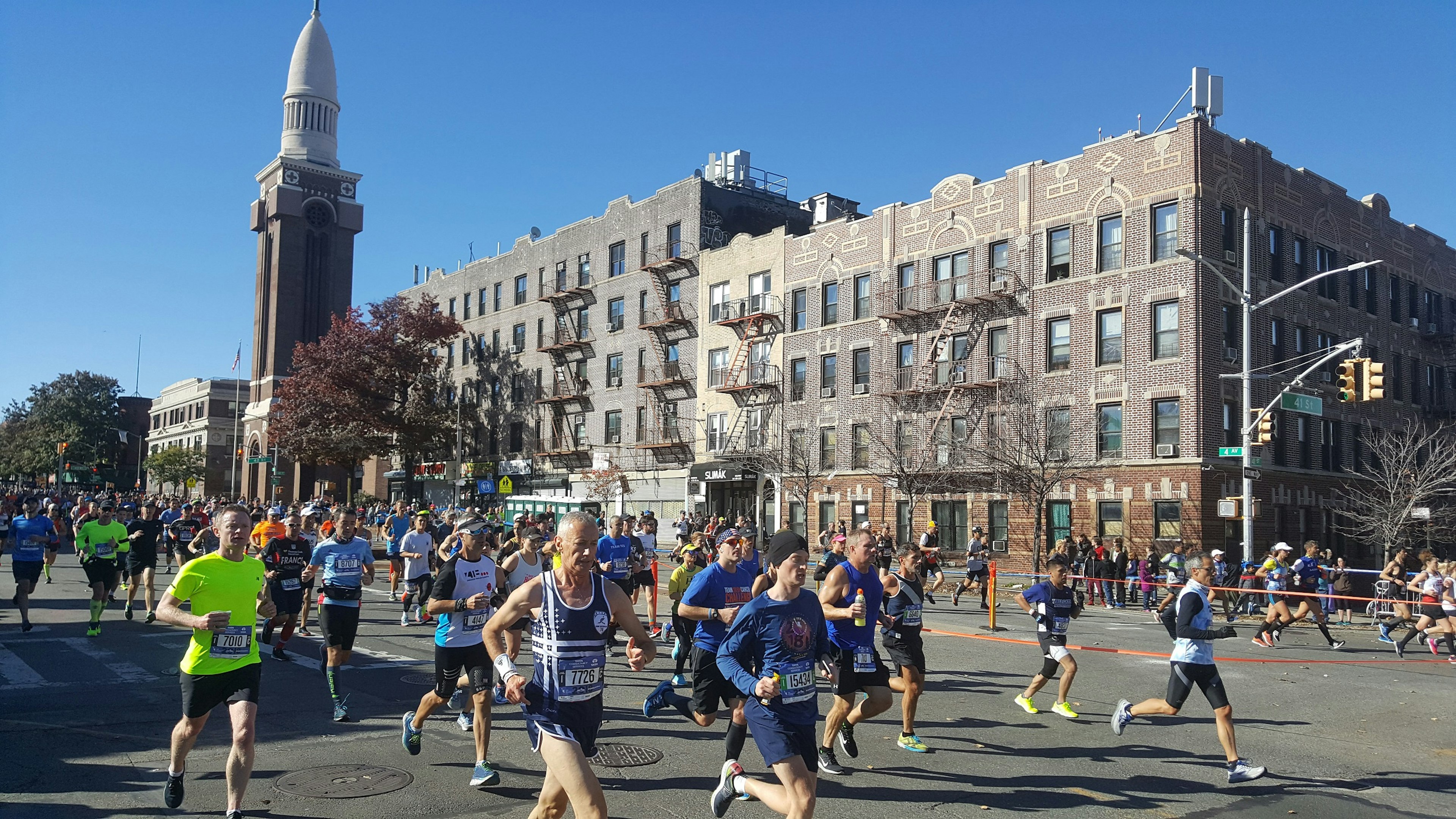 Marathon runners head down a wide-open boulevard in New York City under blue skies.