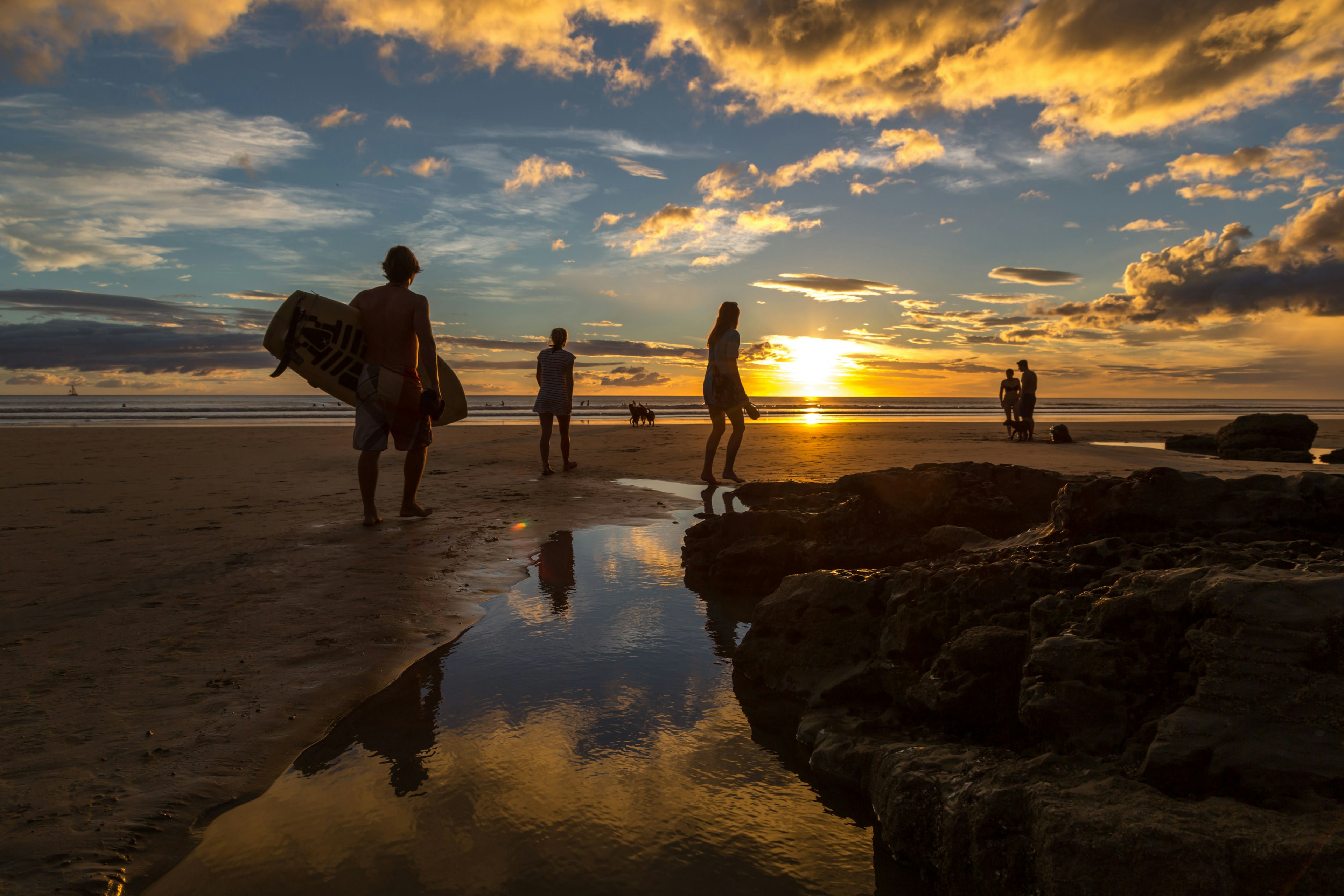 The sun sets on Playa Maderas beach, Nicaragua. A group of people walk across the sand towards the sea, one of which carries a surfboard under his arm.