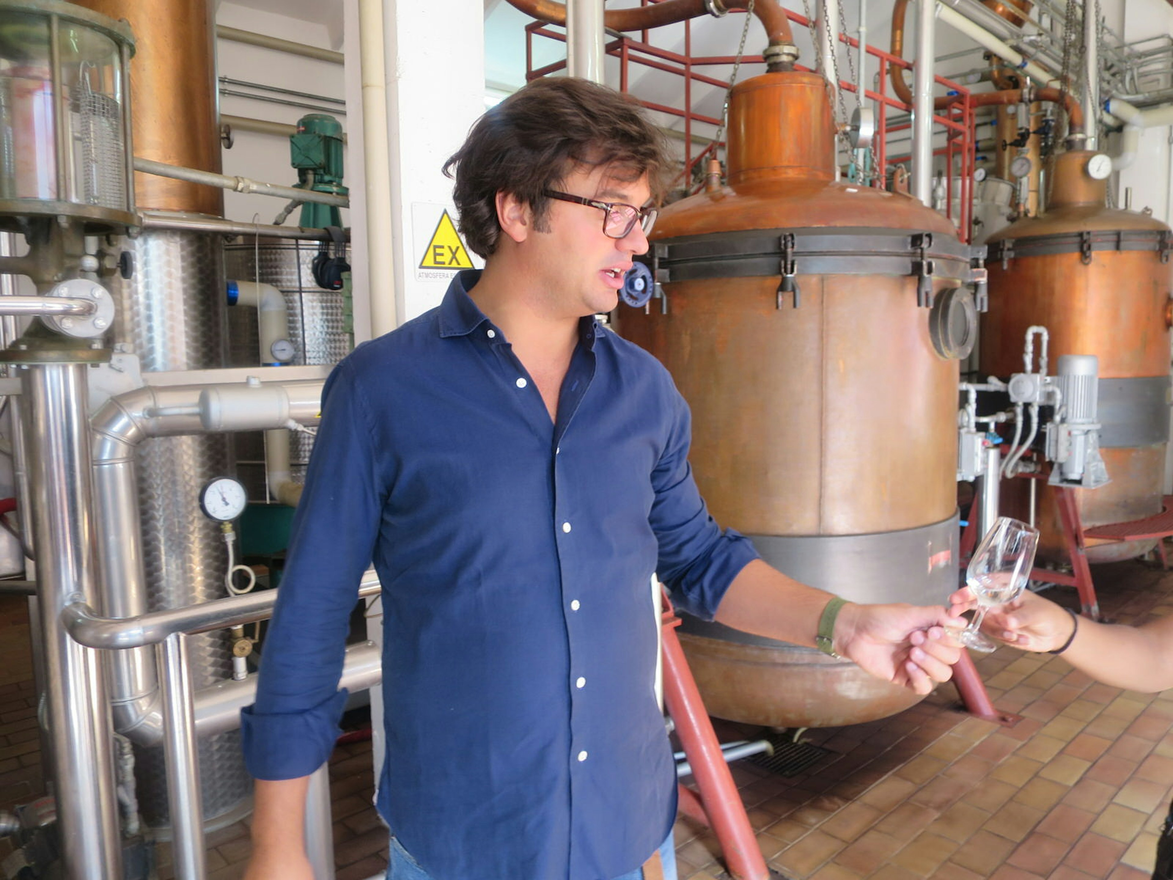 Nicolò Luxardo stands in front of a copper still with a small, full-bodied glass in his hand. He is looking down at the glass and talking.