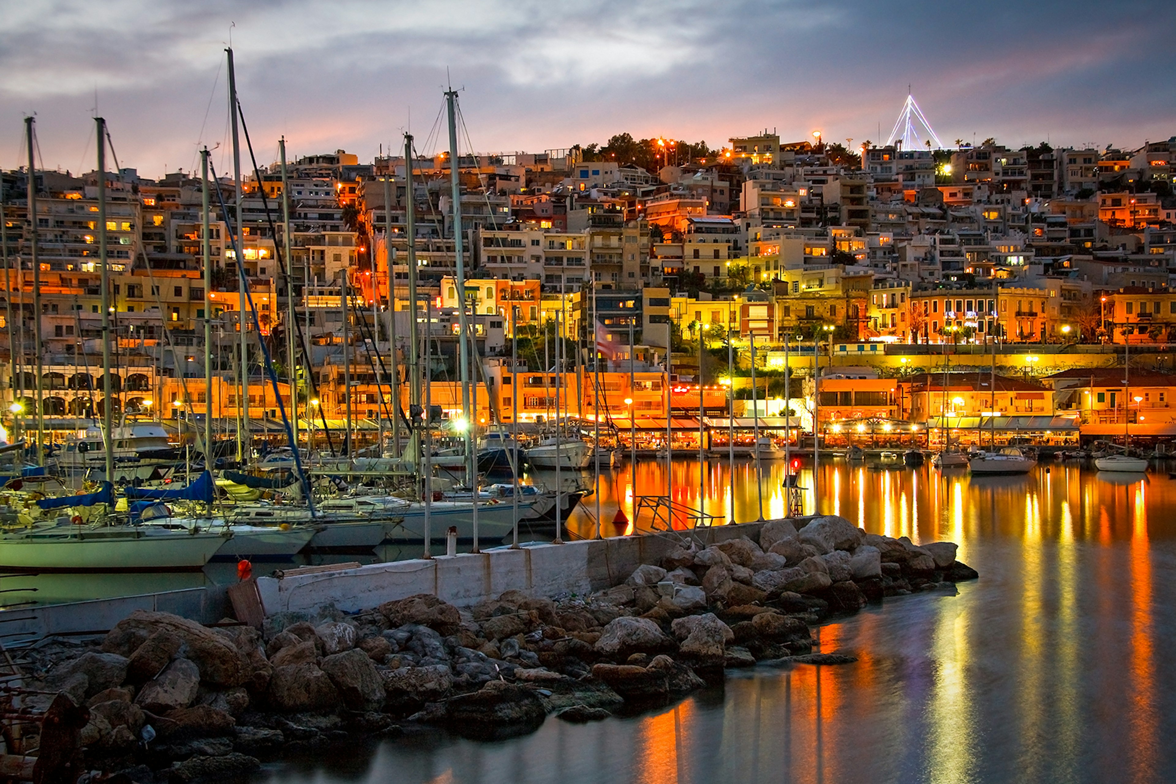 A view looking up at a hillside in Piraeus covered with buildings; the sun is setting and the streetlights are one, giving everything an orange glow. Piraeus, Greece.