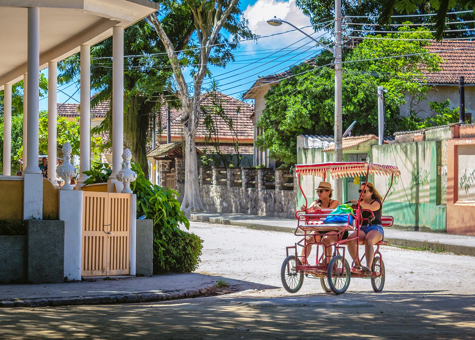 Two women pedal a foot-powered cart through a street lined with pastel-colored buildings on Ilha da Paquetá, Brazil