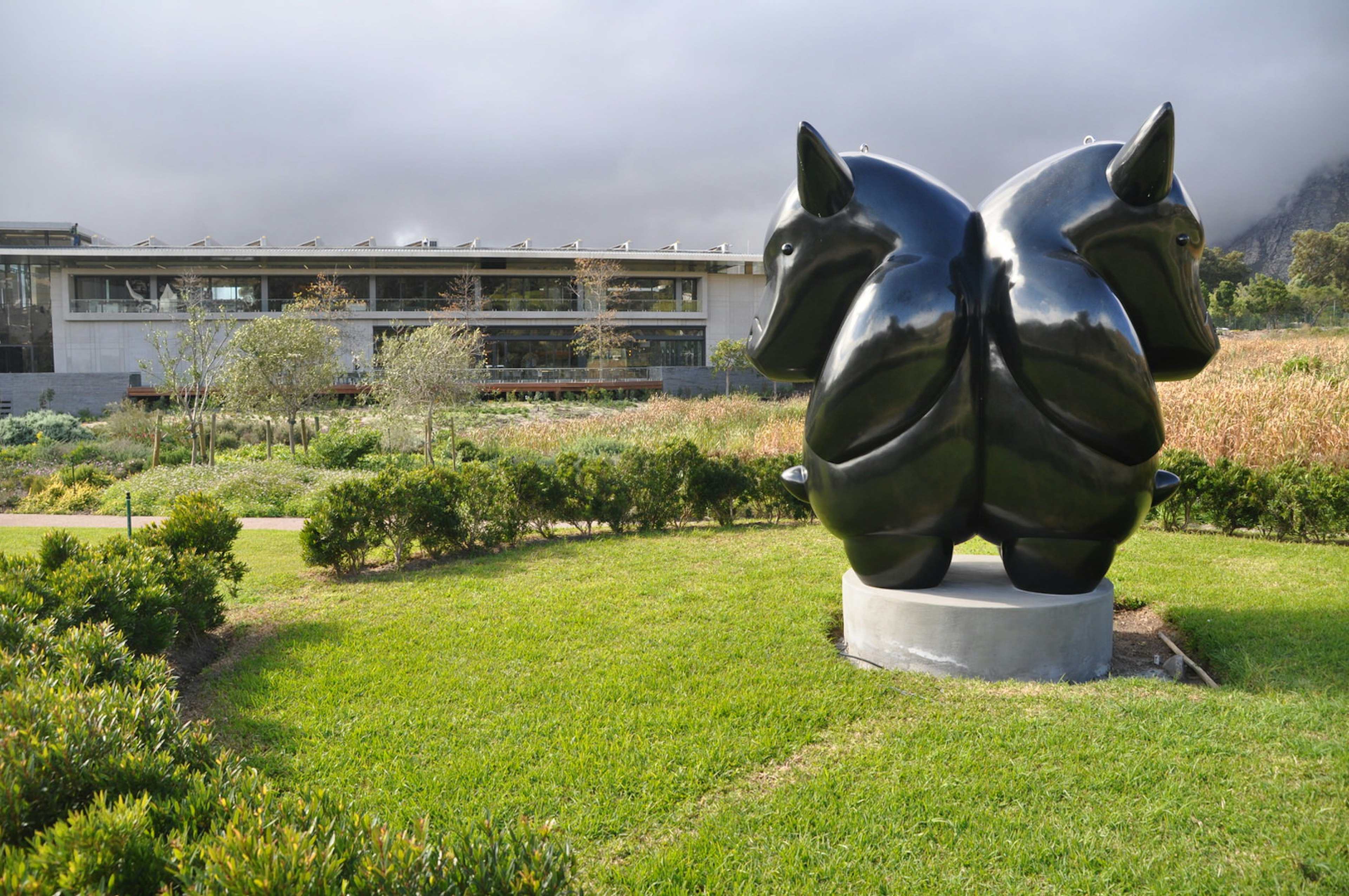 Sitting within a round section of lawn surrounded by a low hedge is a statue of a two-headed hippo-like creature with horns. In the background is the modern building housing the Norval Foundation art museum © Monica Suma / ϰϲʿ¼