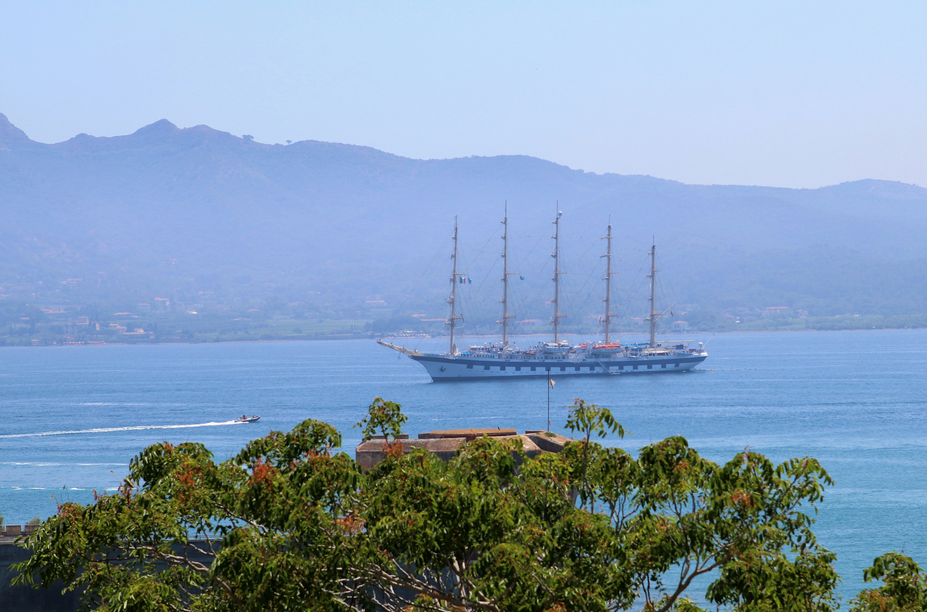 A large schooner glides across a body of water as a smaller speeds past. In the background is a small range of mountains; nude cruise