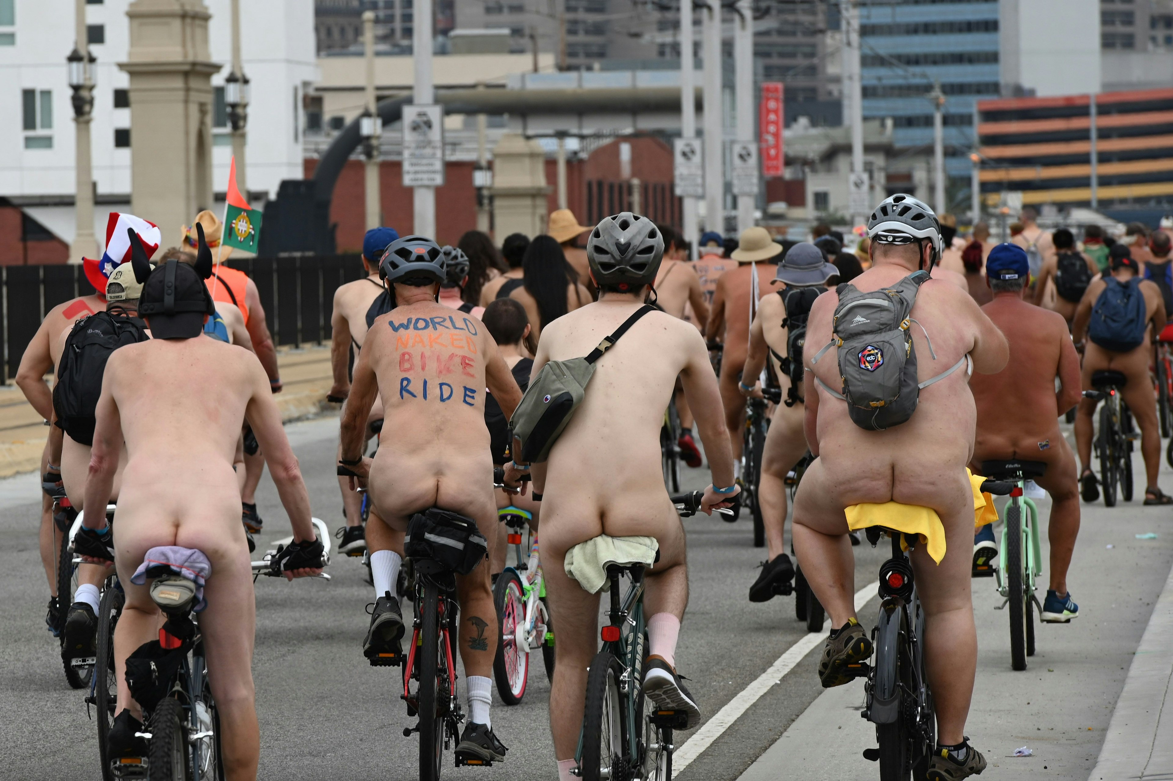 A group of cyclists ride through a city street in the nude. Some are wearing helmets and all are wearing socks and sneakers.