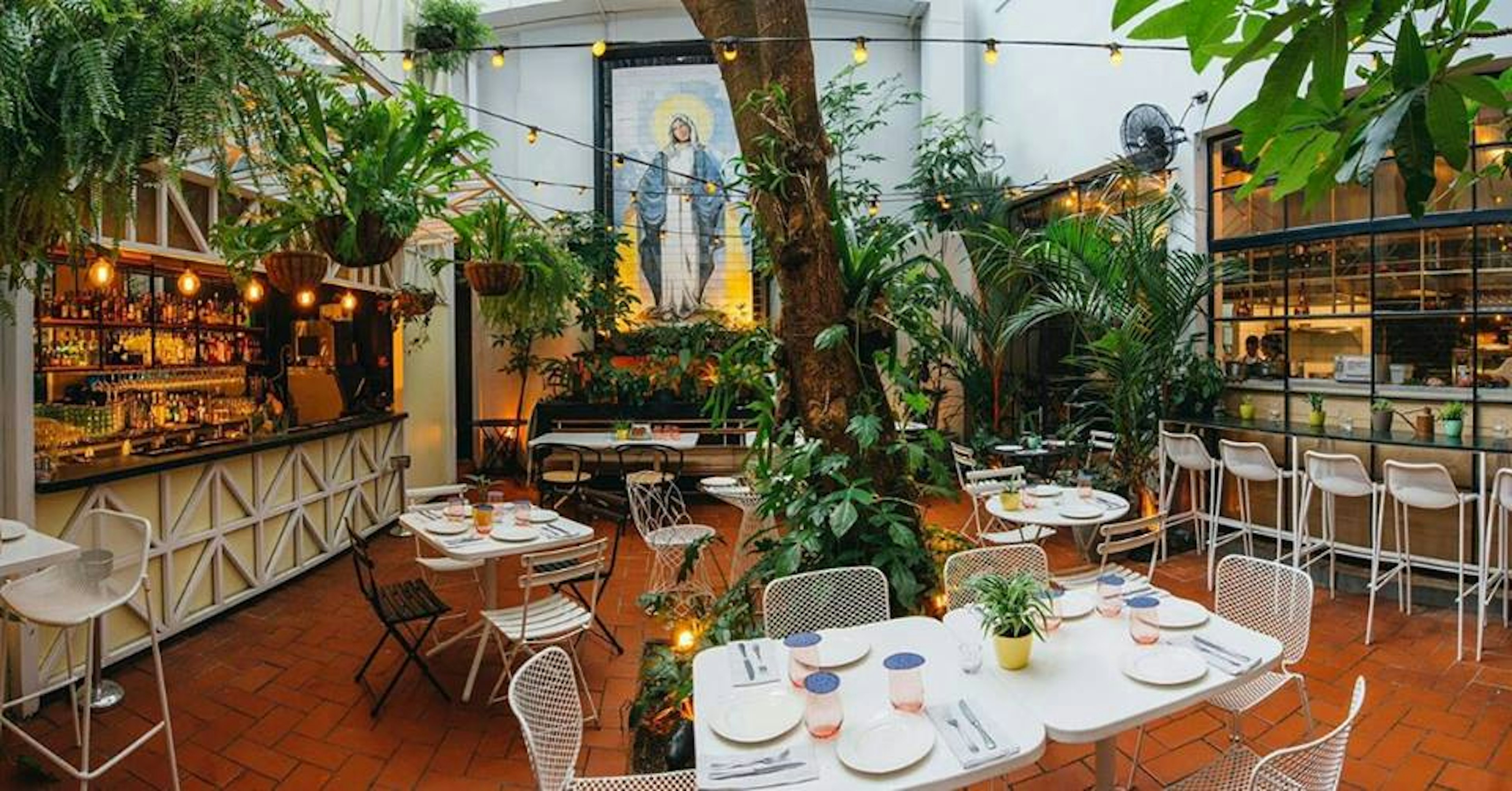 The lush plants, overhead lighting, and large mural of the Virgin Mary create a cozy atmosphere at Ochoymedio Restaurant