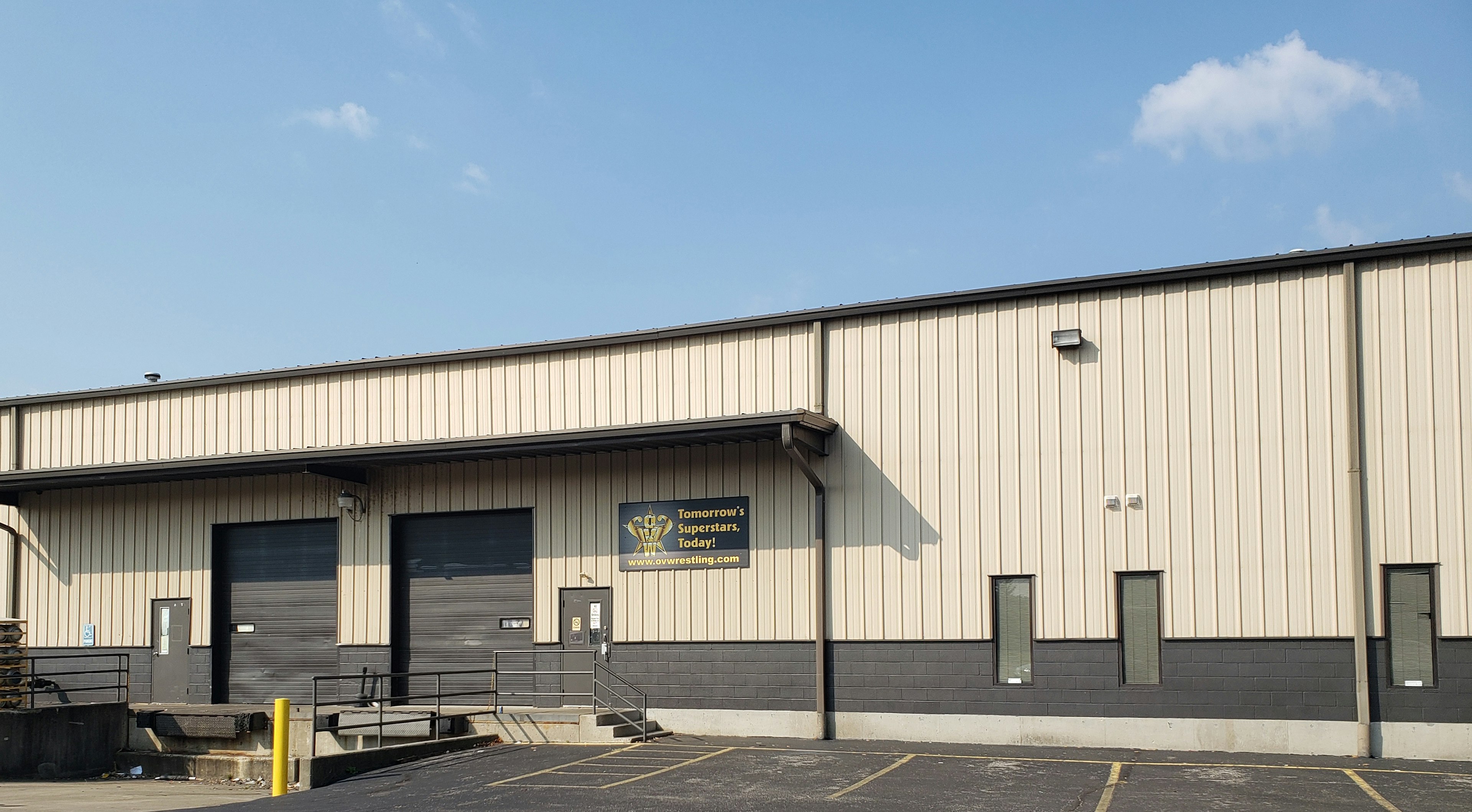 A tan corrugated metal exterior of the Ohio Valley Wrestling trade school has an industrial appearance, complete with two large, dark brown garage doors over loading docks. A black sign by the door with the OVW logo has bright yellow letters that read Tomorrow's Superstars, Today!