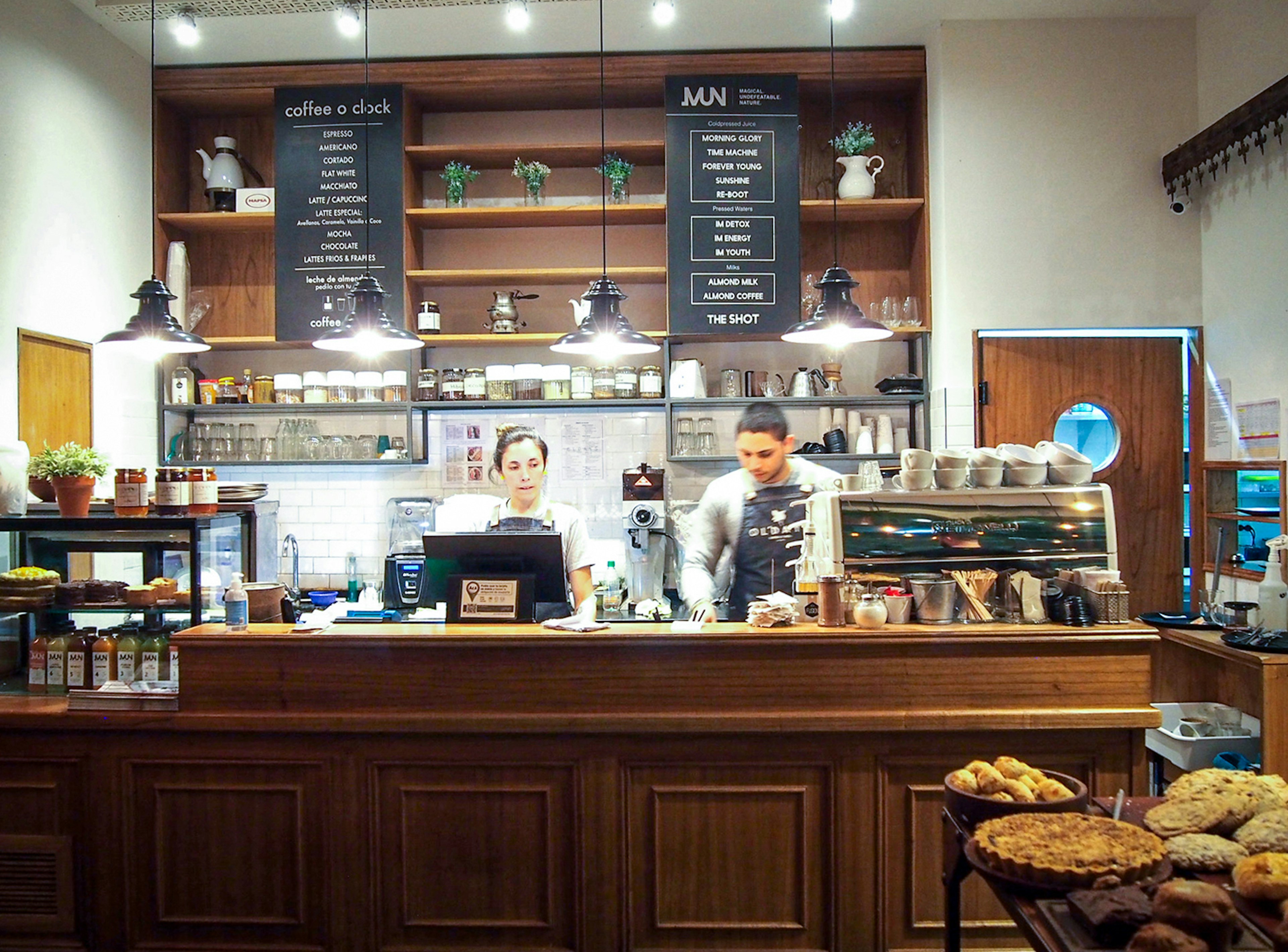 Two people work behind a wooden counter with pies and beverages displayed around them