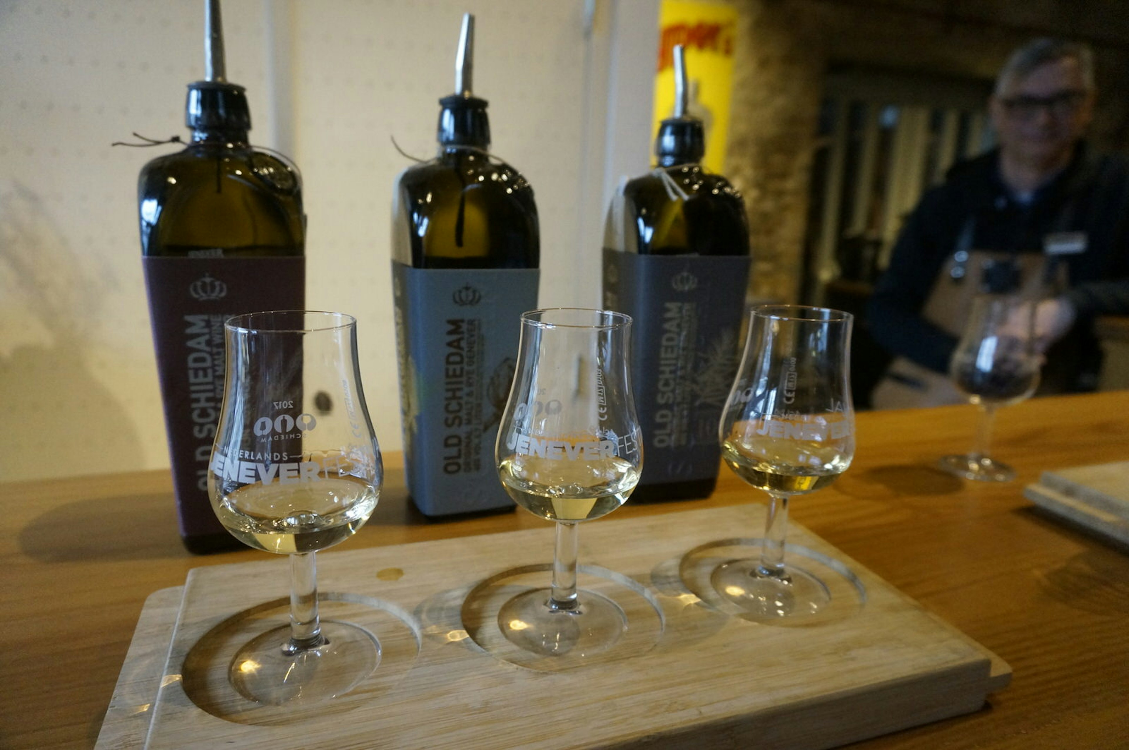 Three tulip-shaped jenever glasses stand on a board on a tabletop with accompanying bottles stood behind them