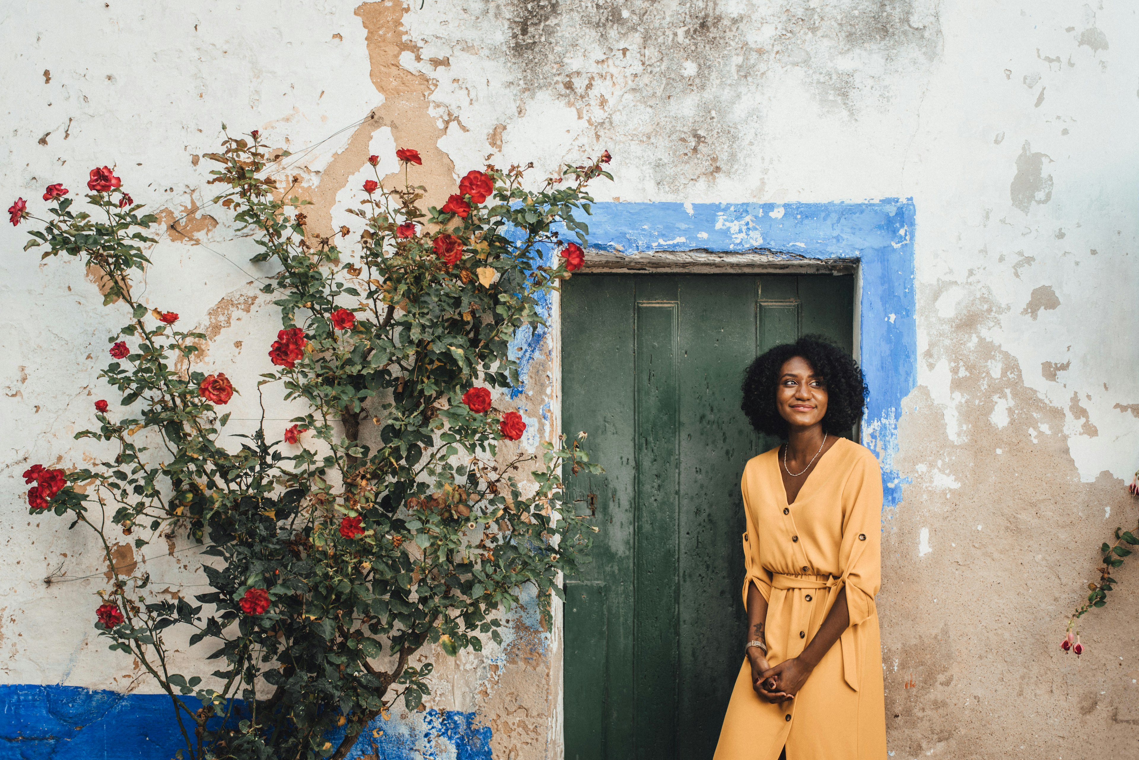 Olivia De Santos stands in front of a white-washed building with a rose plant curving up the wall.