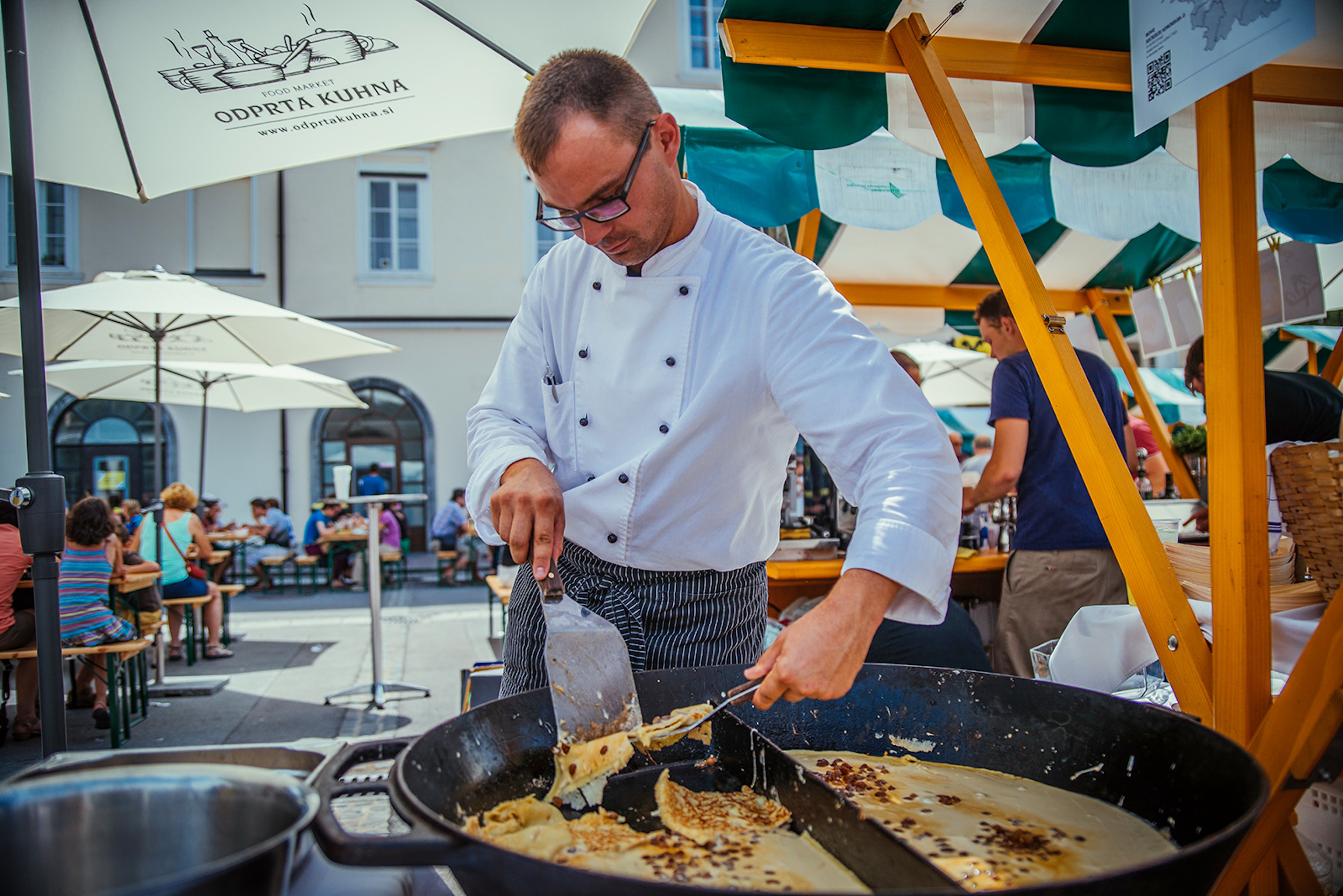 A check cooking in an open-air food market with people eating at umbrella-shaded picnic tables