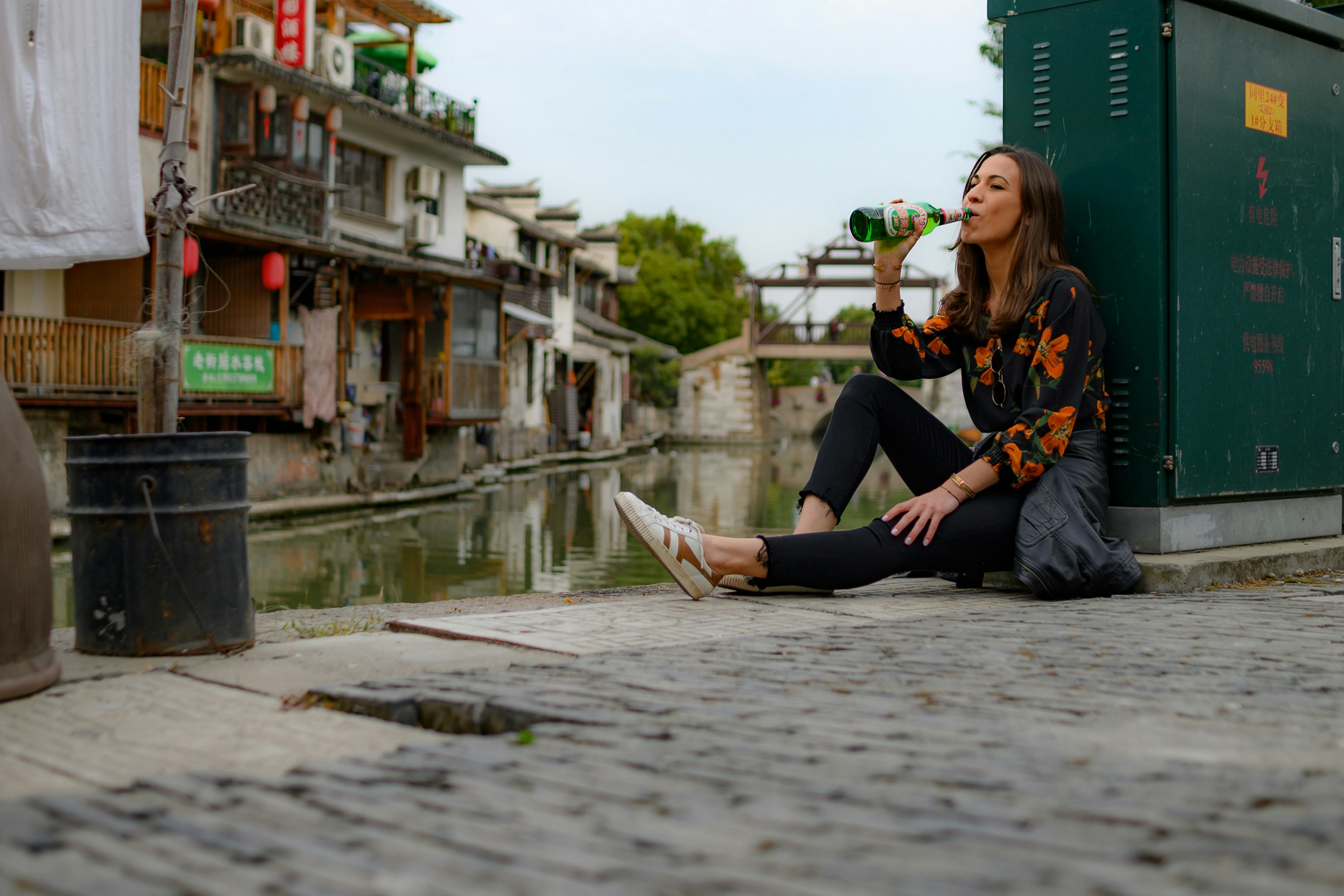 A woman drinks a beer while sitting on the cobbled-street of Suzhou. In the background, one of the city's canals is visible.