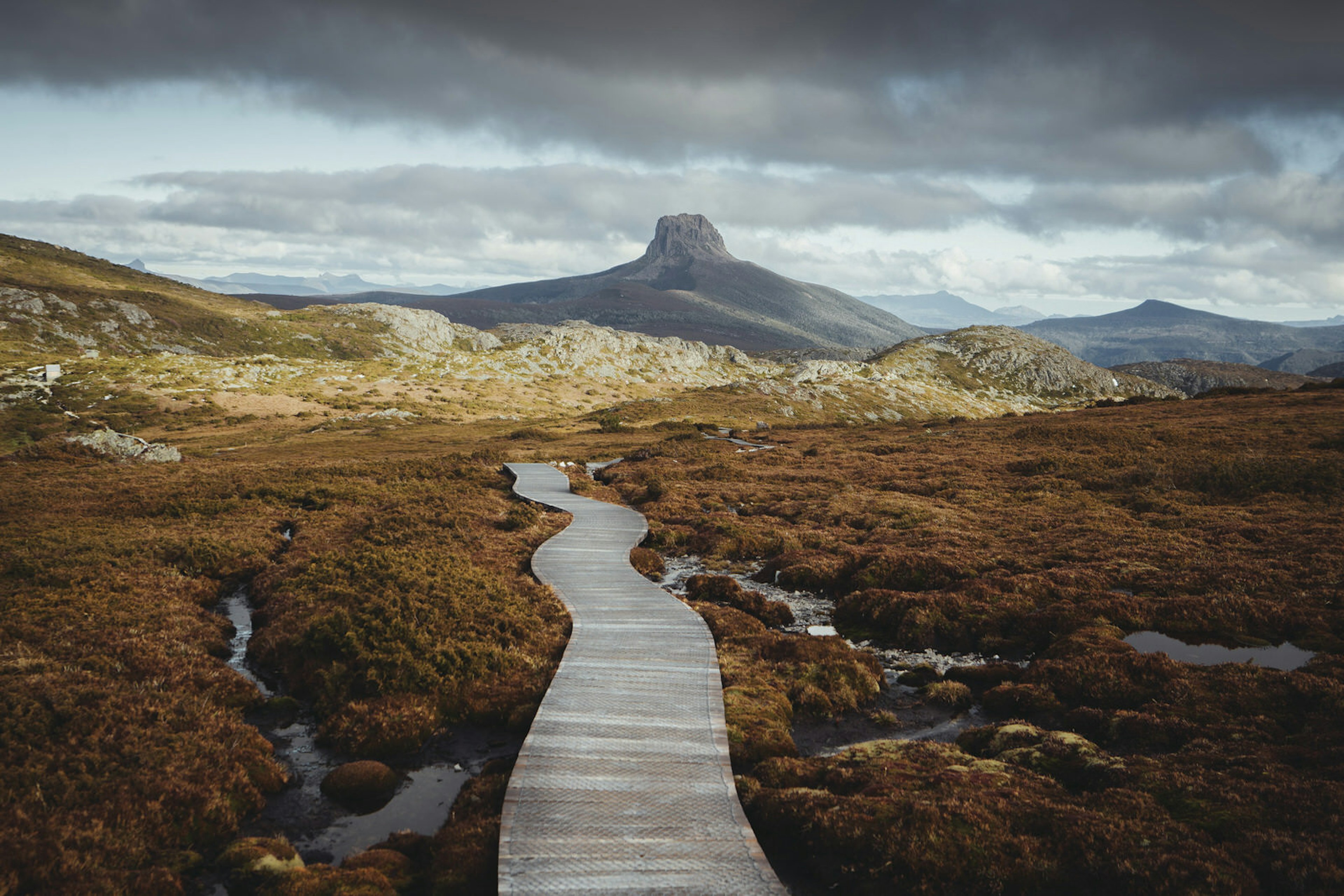 A wooden footpath stretches away from the camera over russet shrubbery and rock pools on Tasmania's Overland Trail. There is a distinctly square rocky peak in the distance underneath an overcast sky