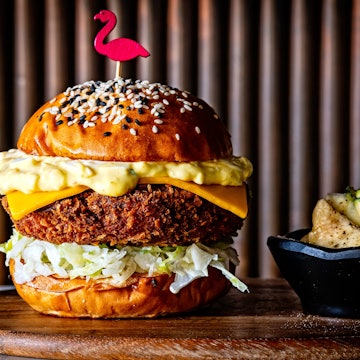 A tall, layered burger with a sesame seed bun and a flamingo toothpick topper sits on a wood plank next to pickles in a small bowl. Shrimp katsue burger from Perry's in Washington DC