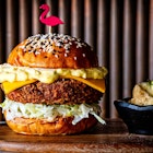 A tall, layered burger with a sesame seed bun and a flamingo toothpick topper sits on a wood plank next to pickles in a small bowl. Shrimp katsue burger from Perry's in Washington DC
