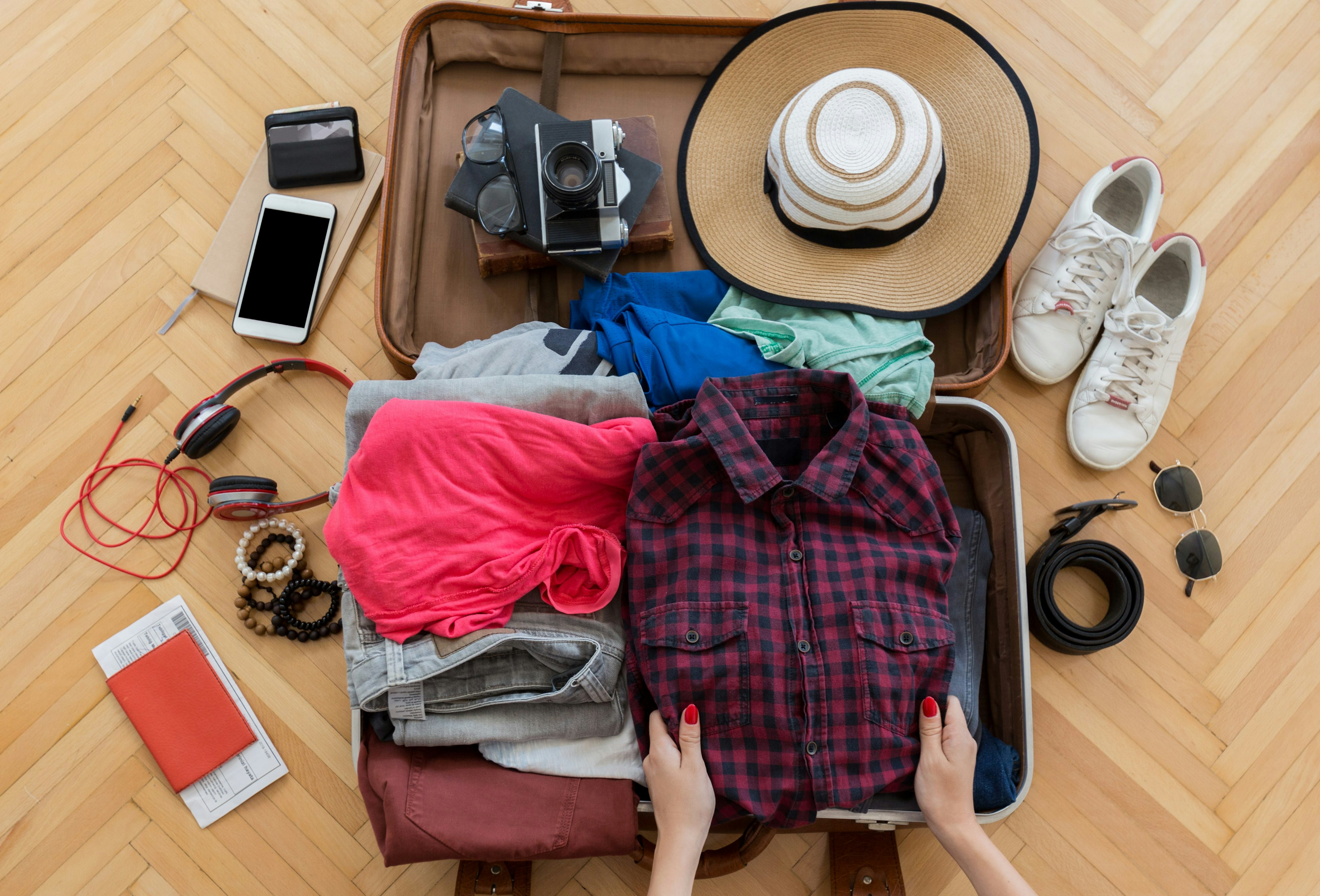 A top-down view of a person packing up a suitcase, which is open on the table. The suitcase is filled with clothing and around it are travel items like a phone, headphones and a passport.