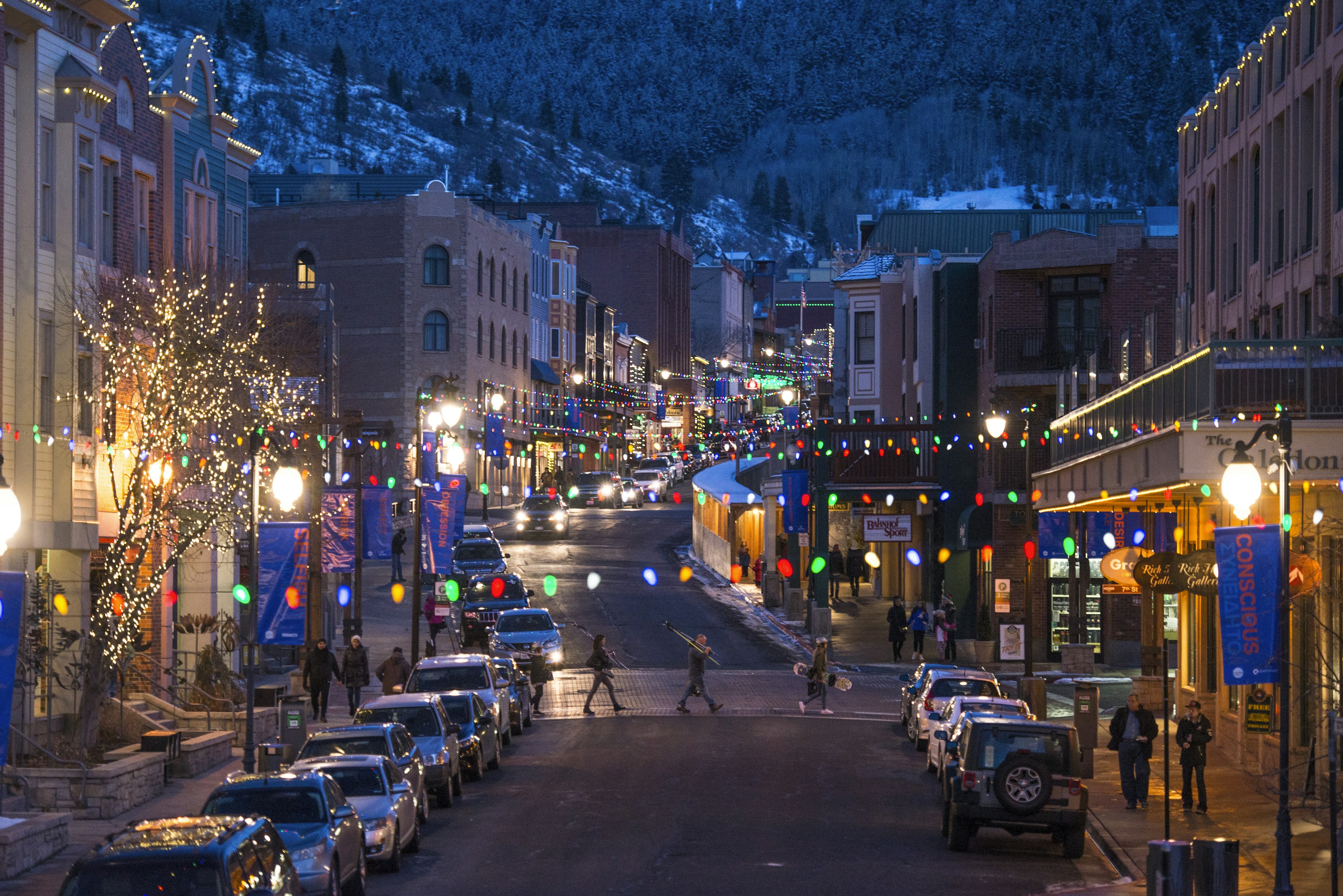 A group of people walk across a cobble-stoned street holding ski gear during the evening in a brightly lit downtown Park City. There are a pair of people each walking downhill on opposite sides of the street; bachelorette party