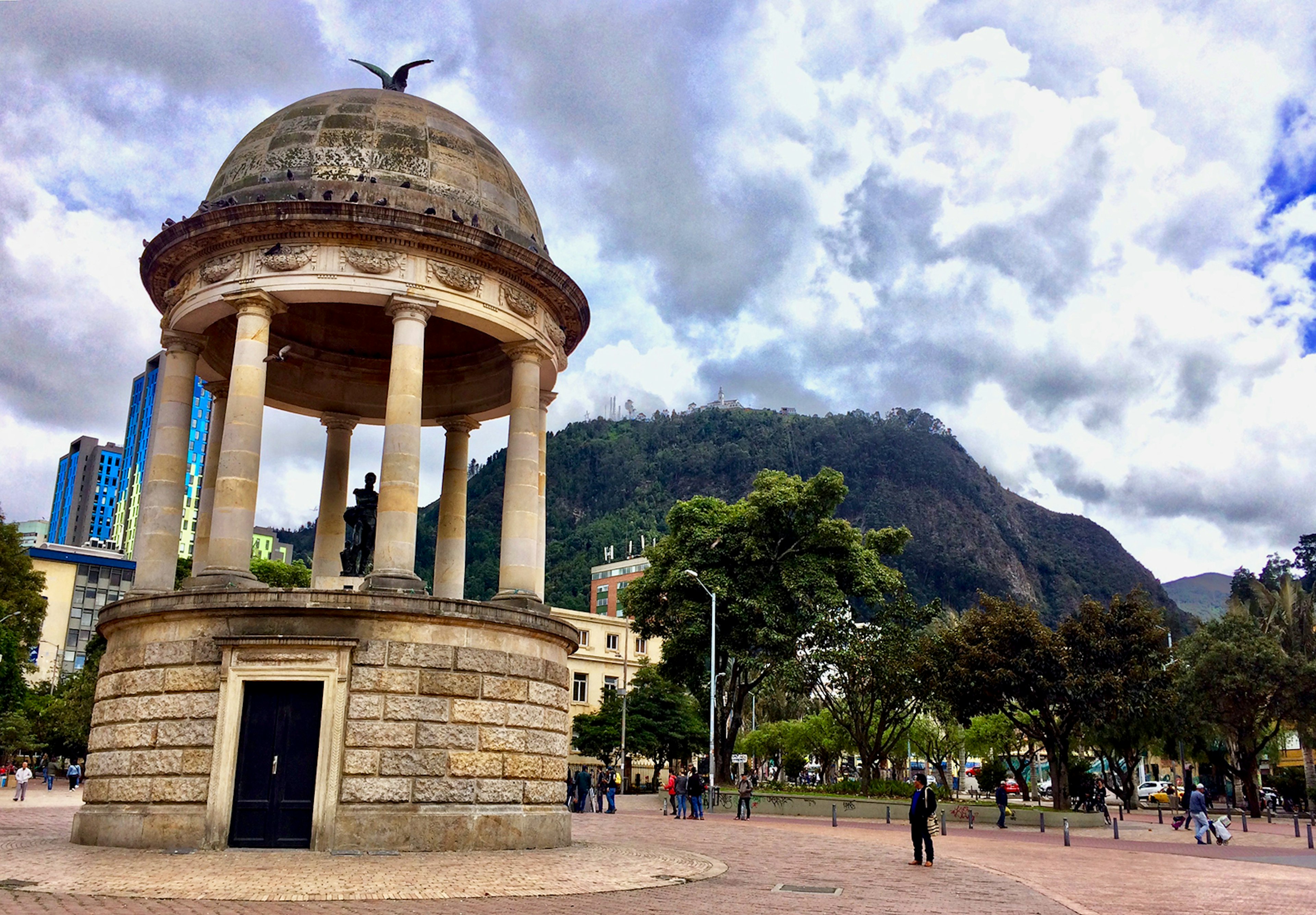 A stone gazebo stands in the middle of a bricked plaza, with green hills in the background