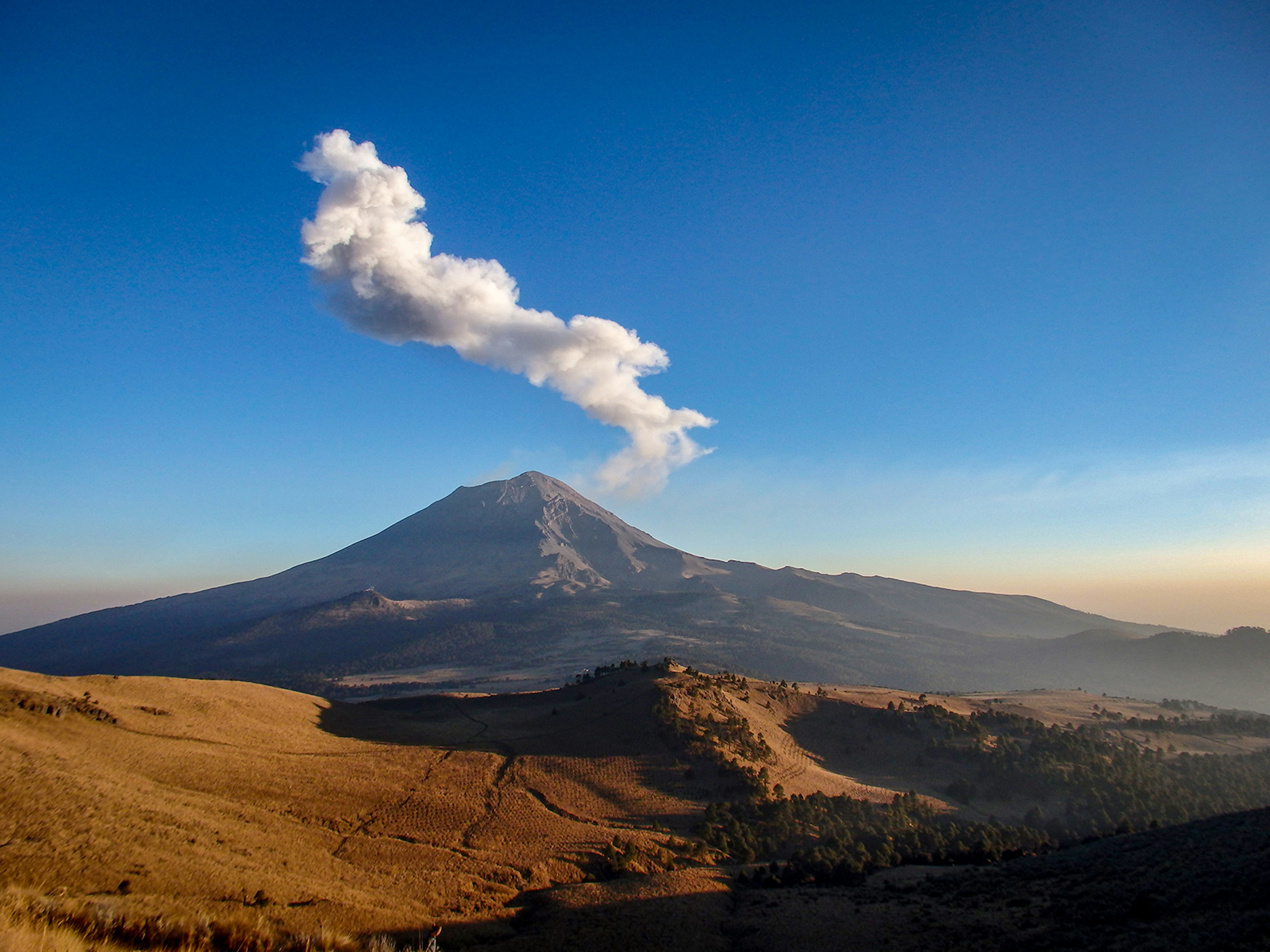 A view of a copper-colored valley stretching toward a volcanic peak with steam coming out in Central Mexico