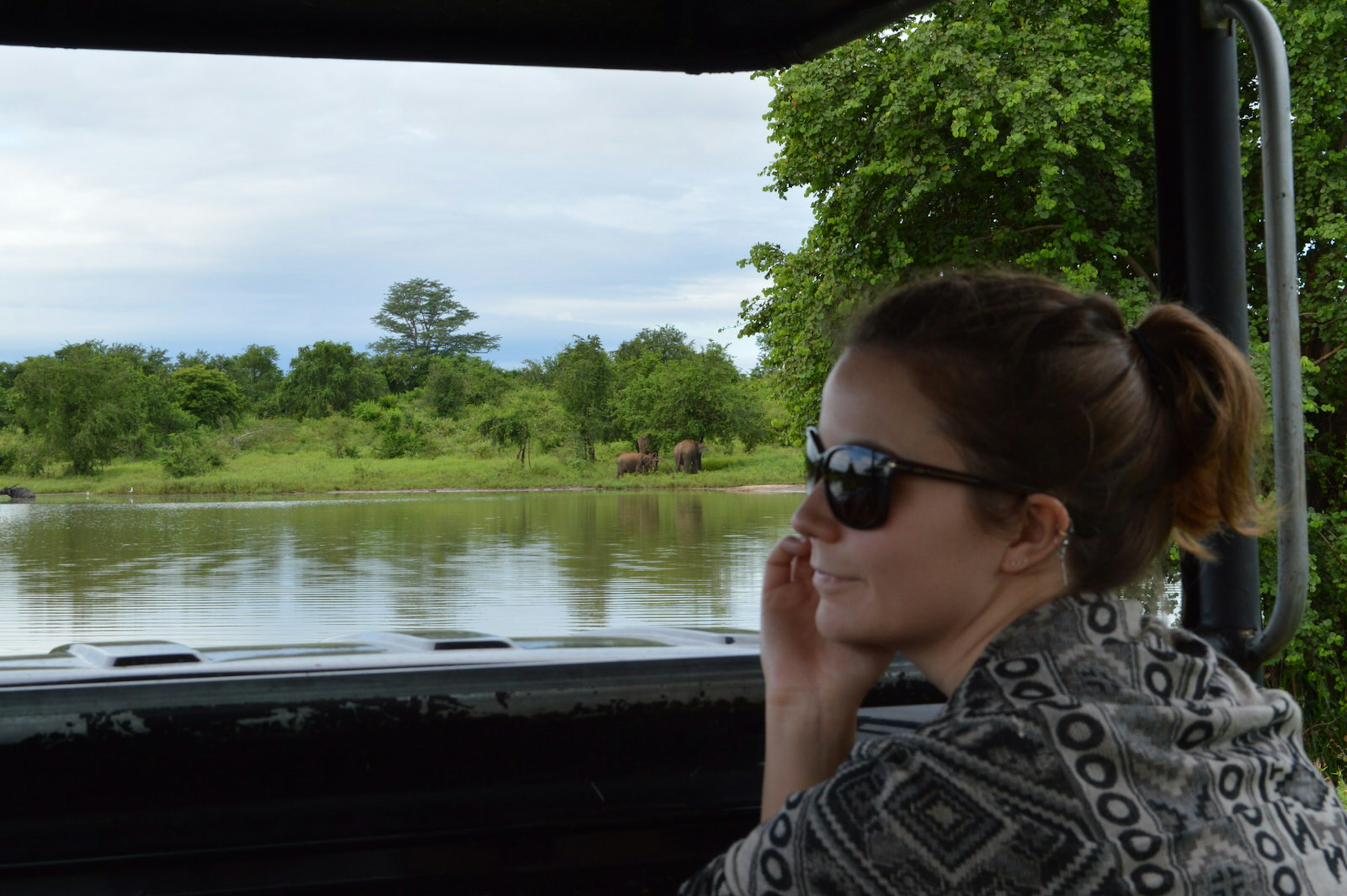 A woman wearing sunglasses sits in a jeep looking out over a lake in Udawalawe, Sri Lanka. There is a small group of elephants on the other side of the lake; travel and grief.